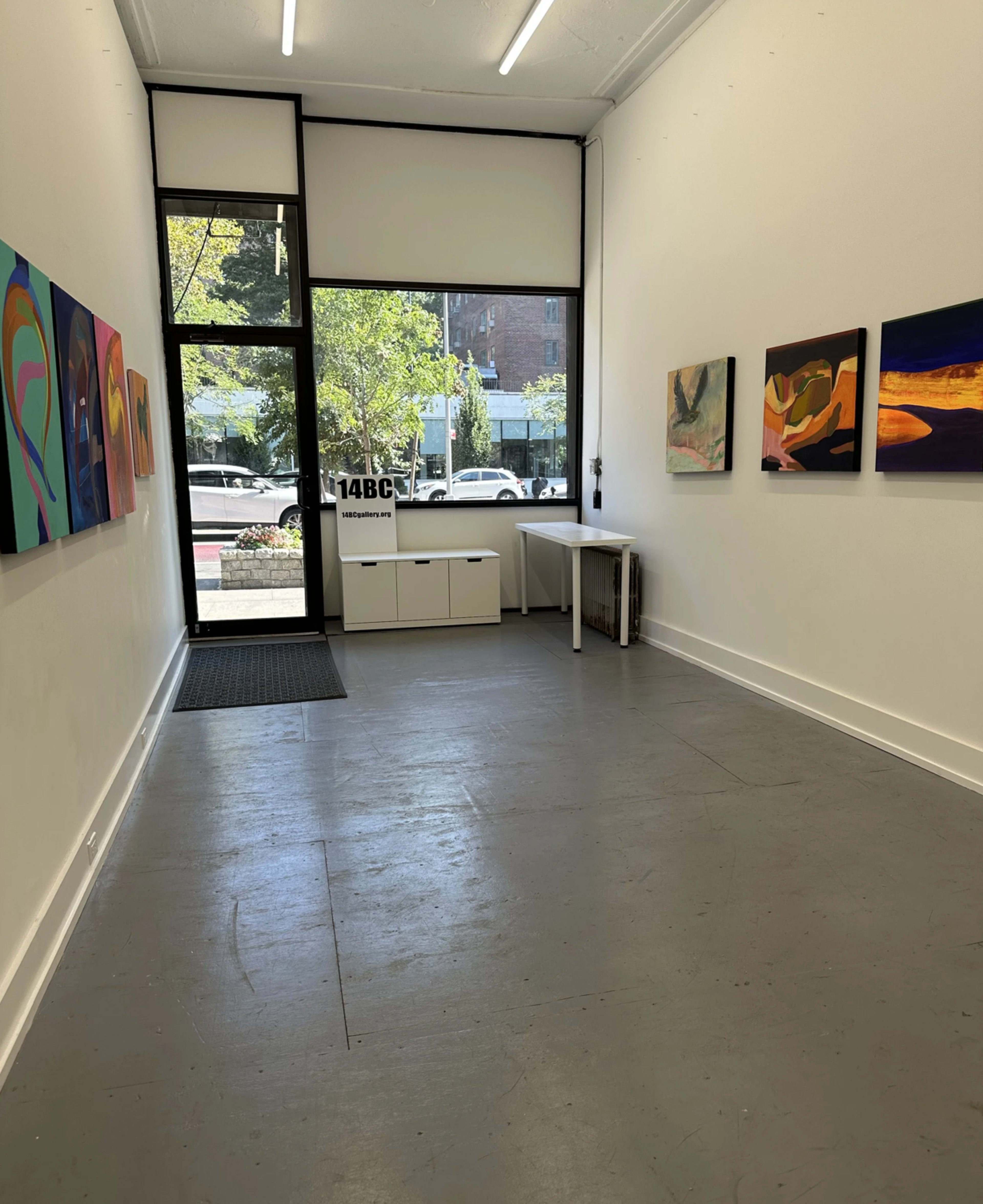 The image shows a spacious art gallery with brightly colored paintings displayed along the walls and a small table near the entrance.