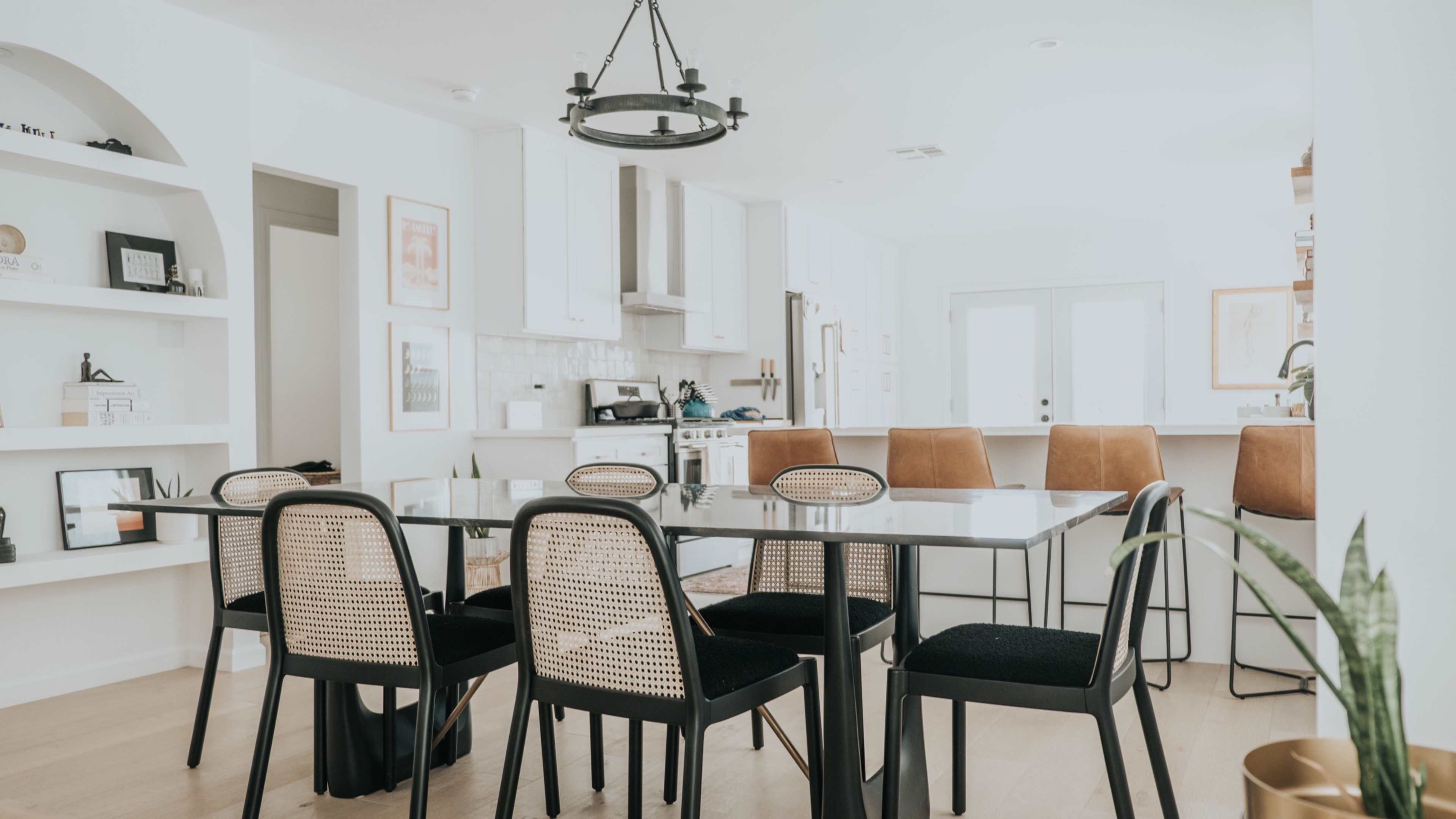 A modern dining area features a glass table surrounded by black and tan chairs, with a kitchen visible in the background.