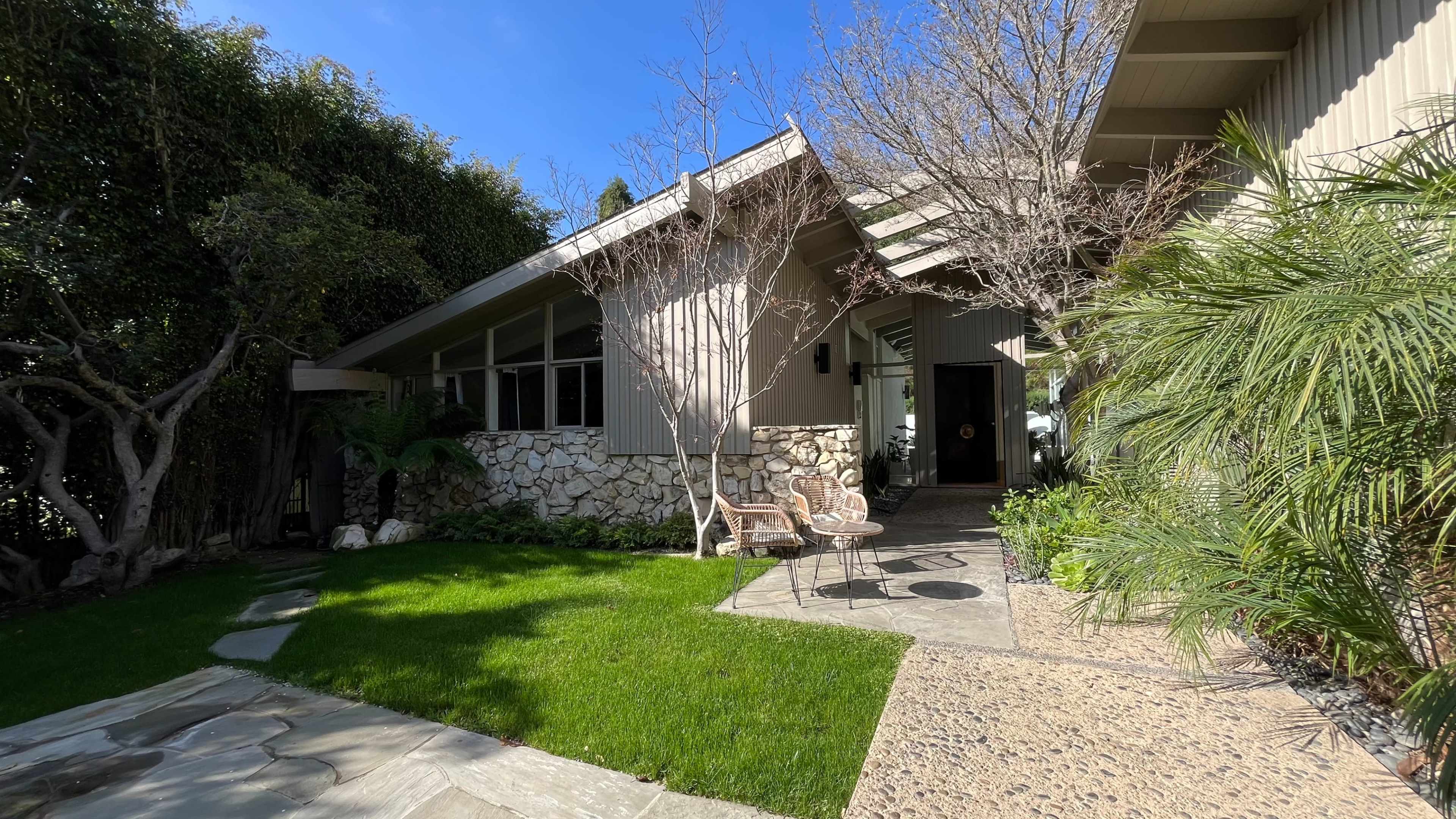 The image shows a modern house with a sloped roof, surrounded by greenery and featuring a stone accent wall and a pathway leading to the entrance.