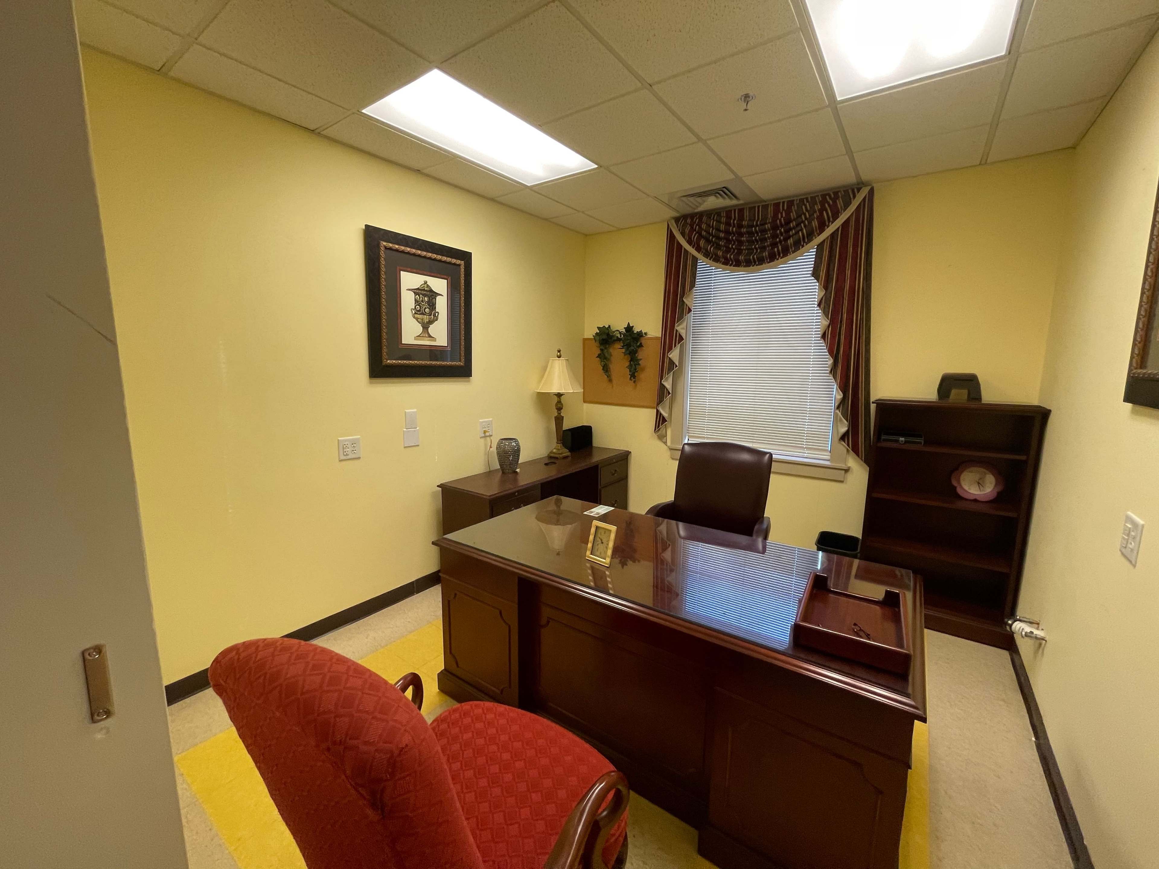 An office with a wooden desk, two chairs, a lamp, and a window covered by curtains, all set against yellow walls.