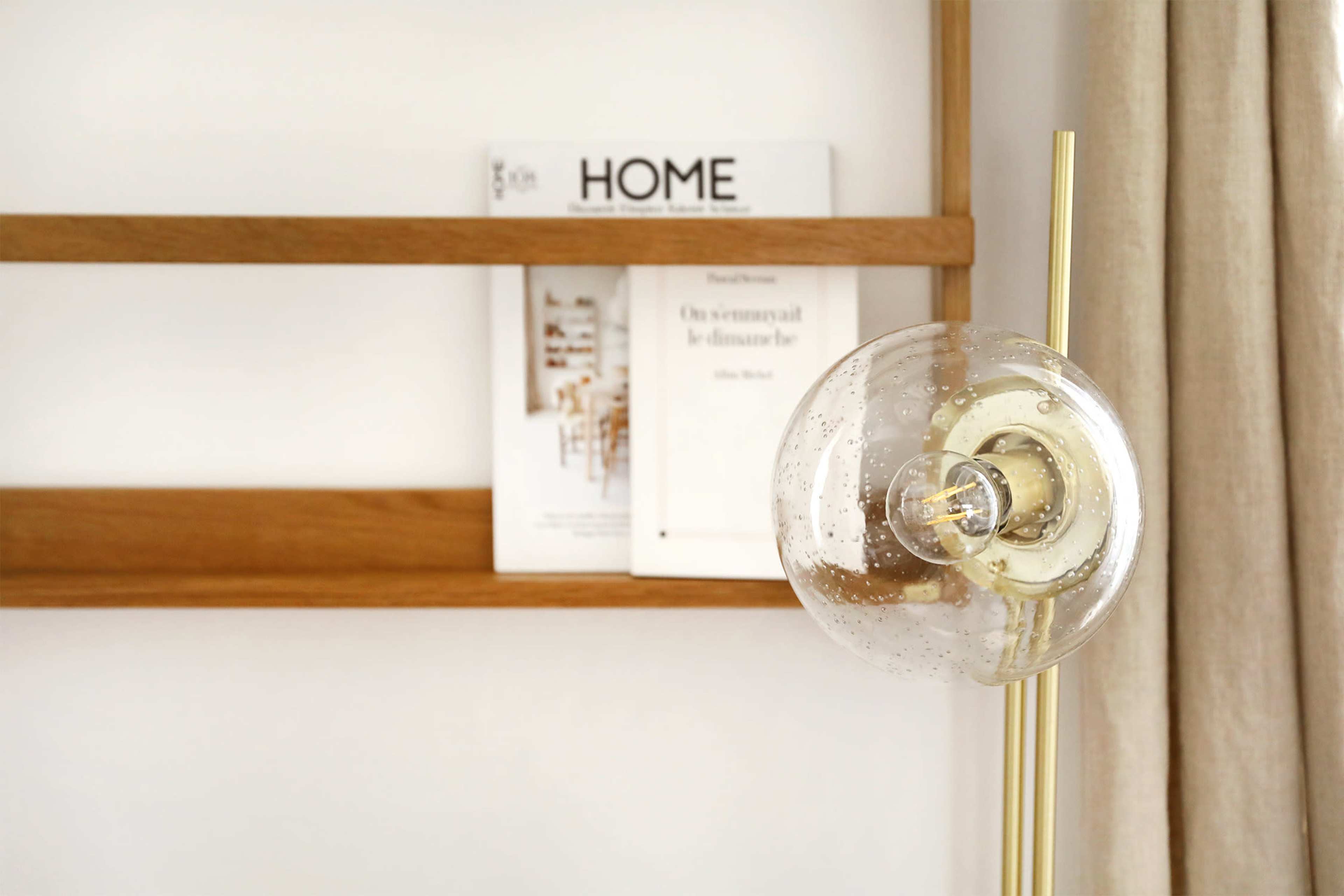 A clear glass lamp with a gold base stands in front of a wooden shelf displaying a magazine titled "HOME."
