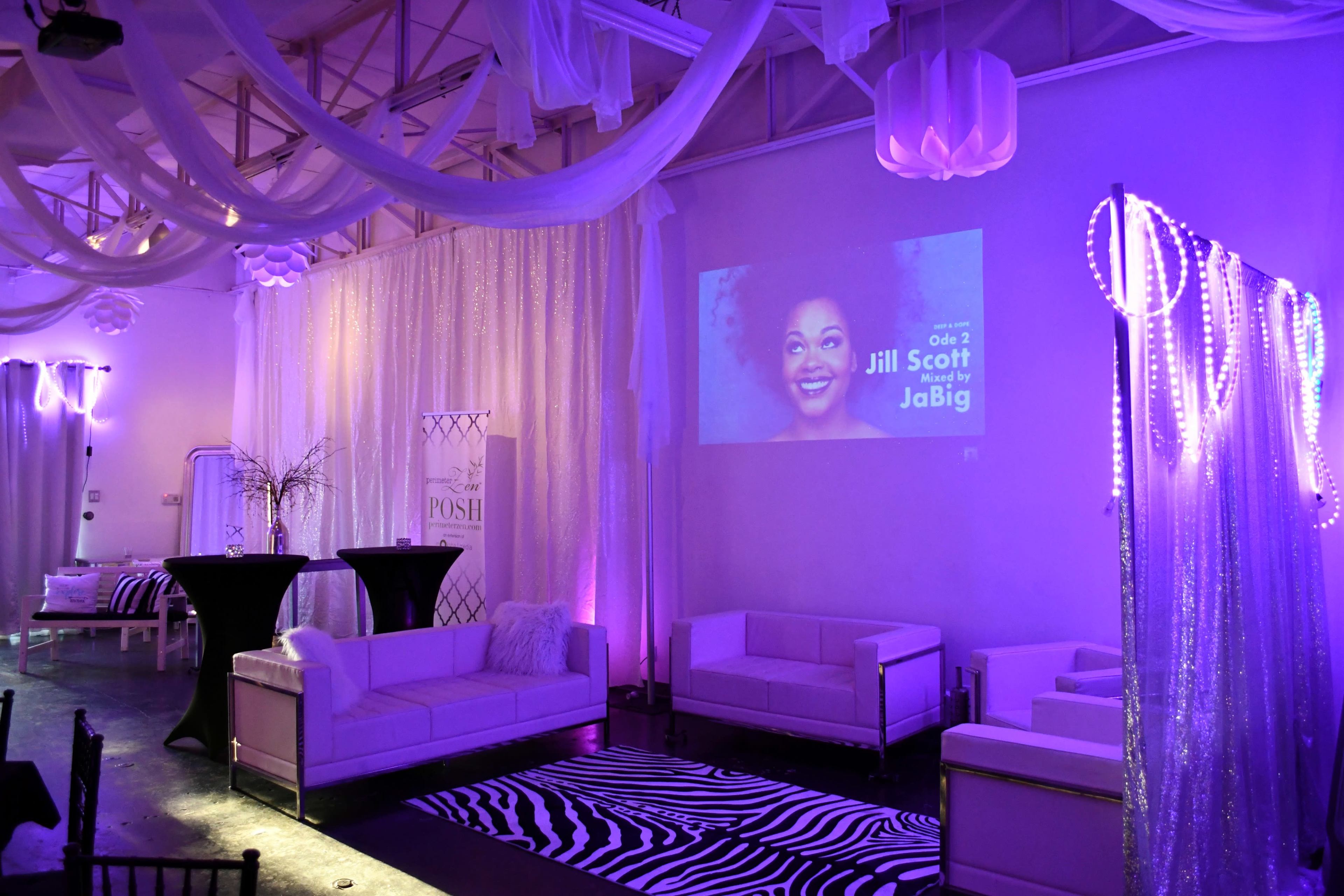 The image shows a stylish event space with white sofas, a zebra-striped rug, and a projector displaying a video, all illuminated by purple lighting.
