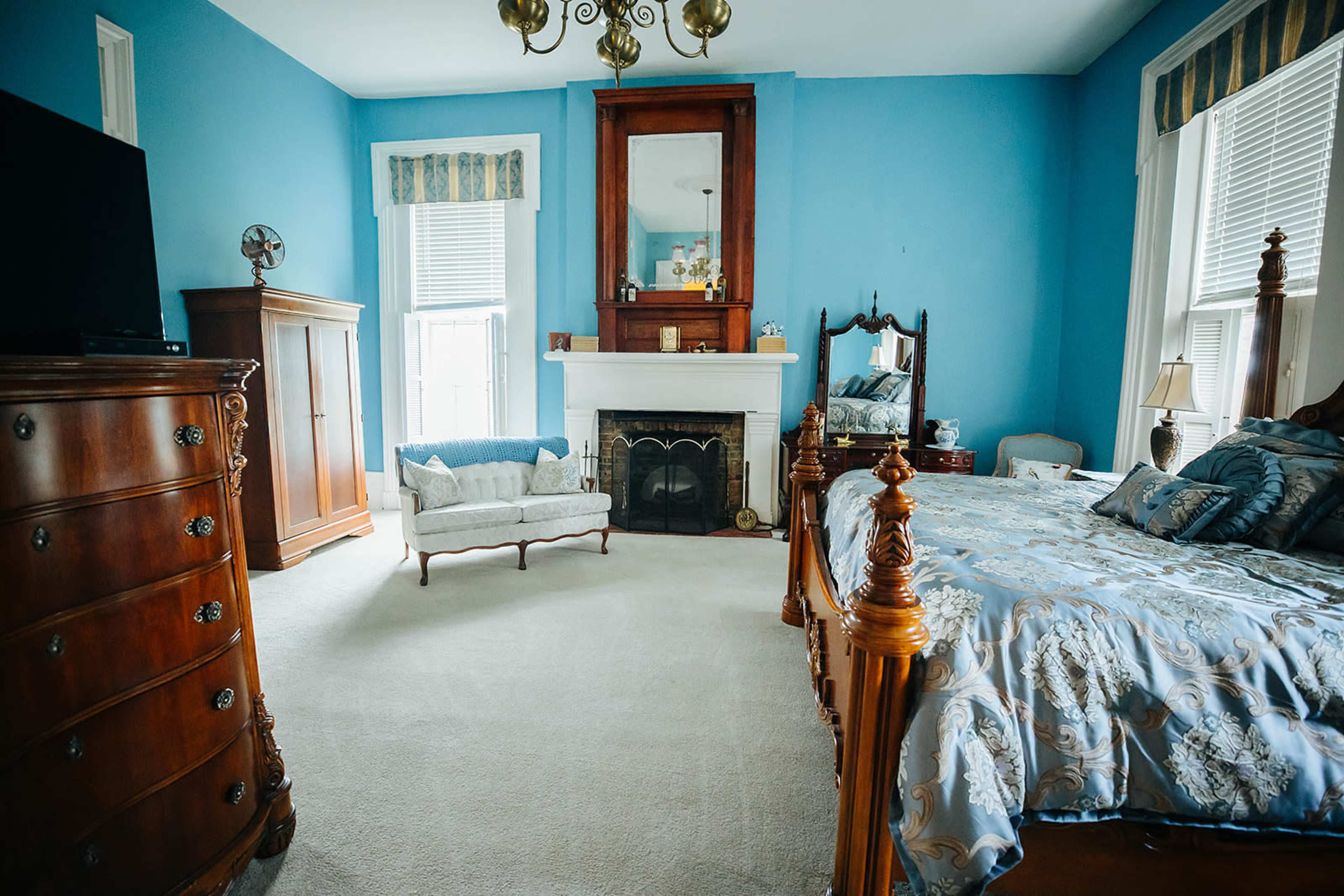 A brightly colored blue bedroom featuring a wooden bed, a fireplace, a dresser, and a small seating area.