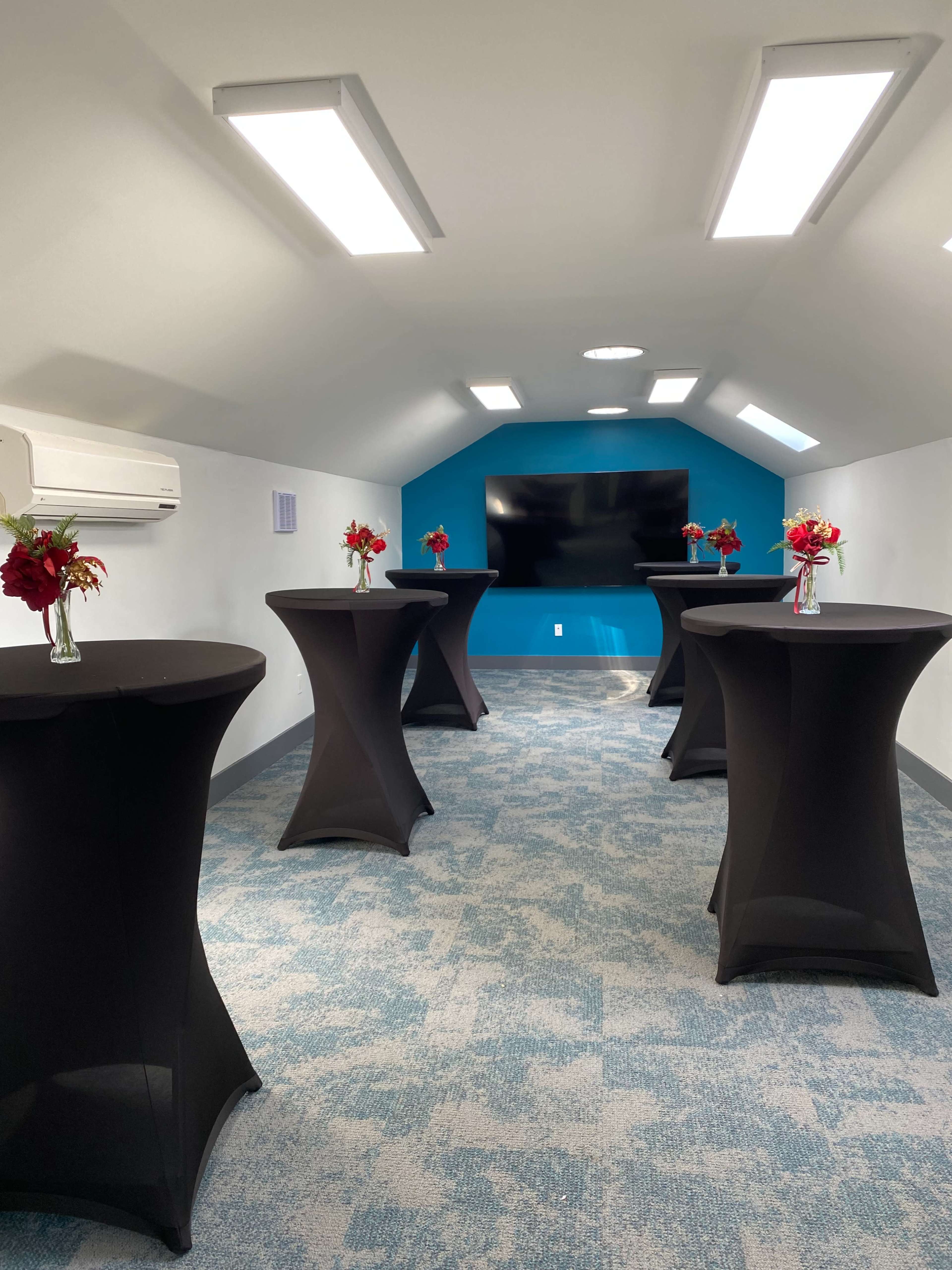 The room features five black cocktail tables with floral centerpieces arranged on a patterned blue carpet beneath bright overhead lights.