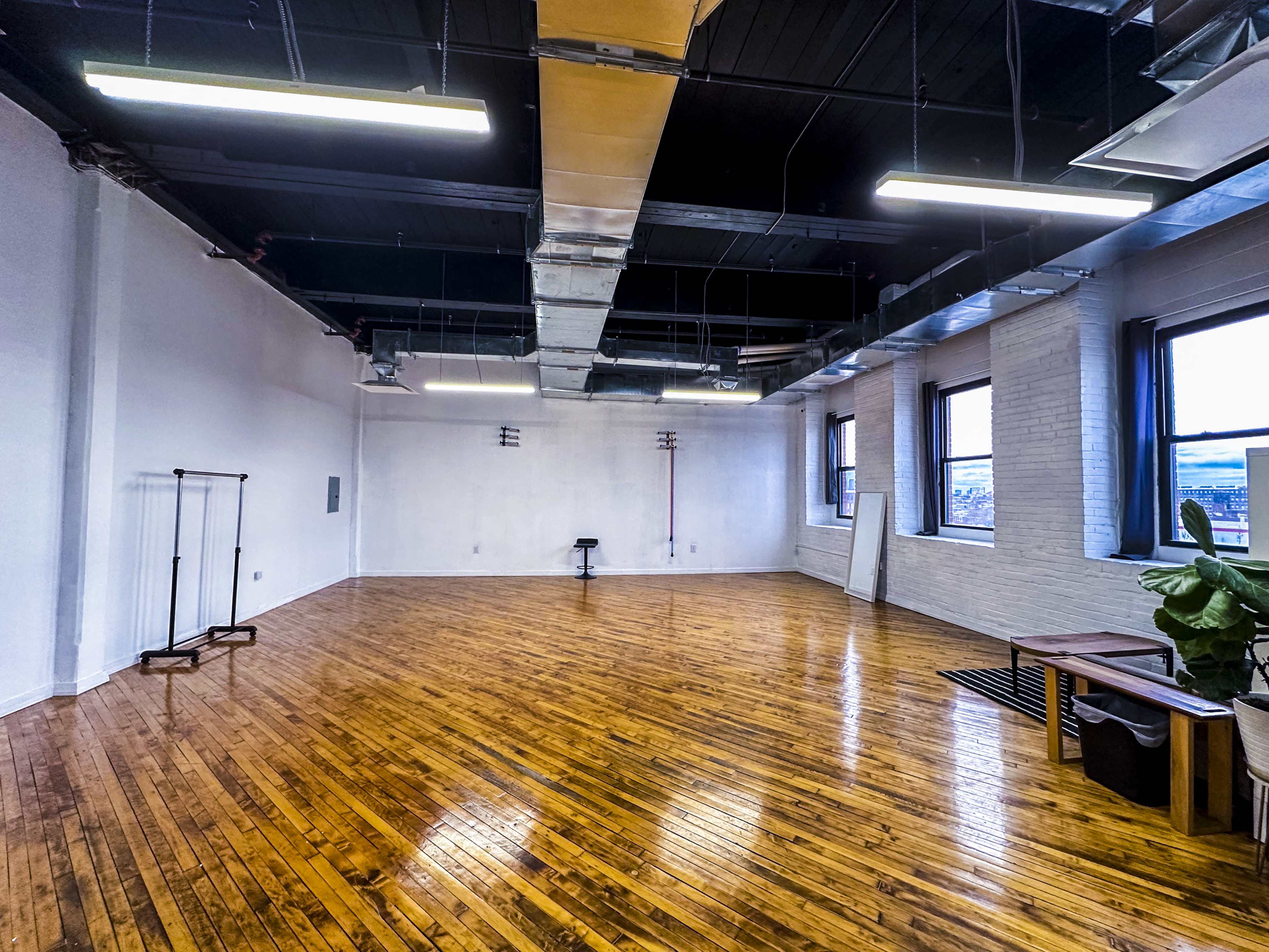 An empty room with hardwood floors, large windows, and industrial-style ceilings, featuring minimal furnishings like a clothing rack and a stool.