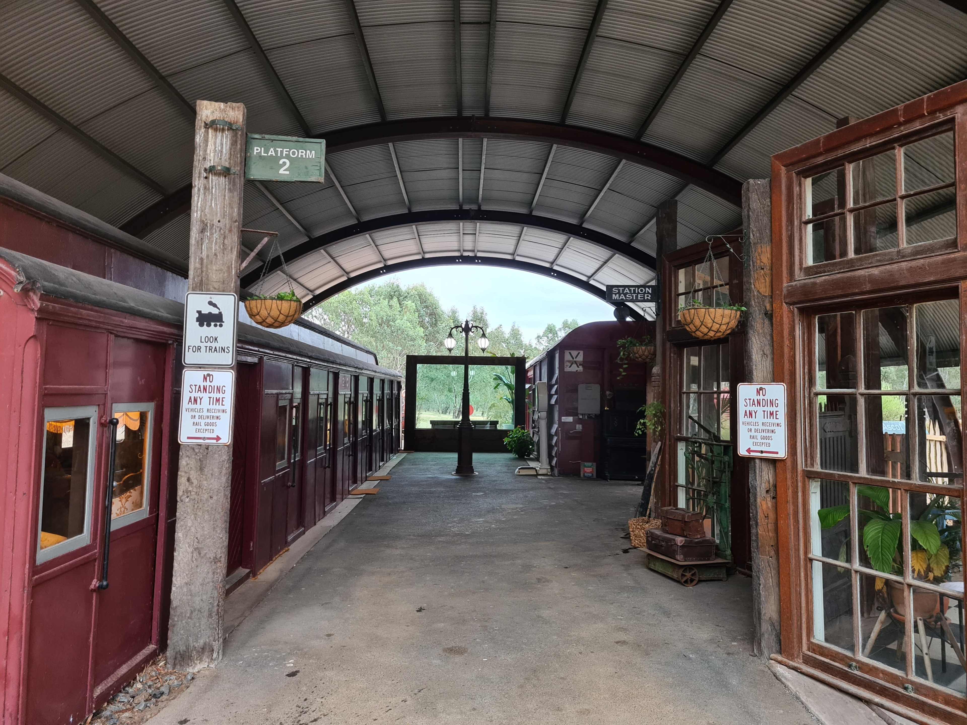 The image shows an open-air train station with wooden stalls and signs indicating the platform and standing area.