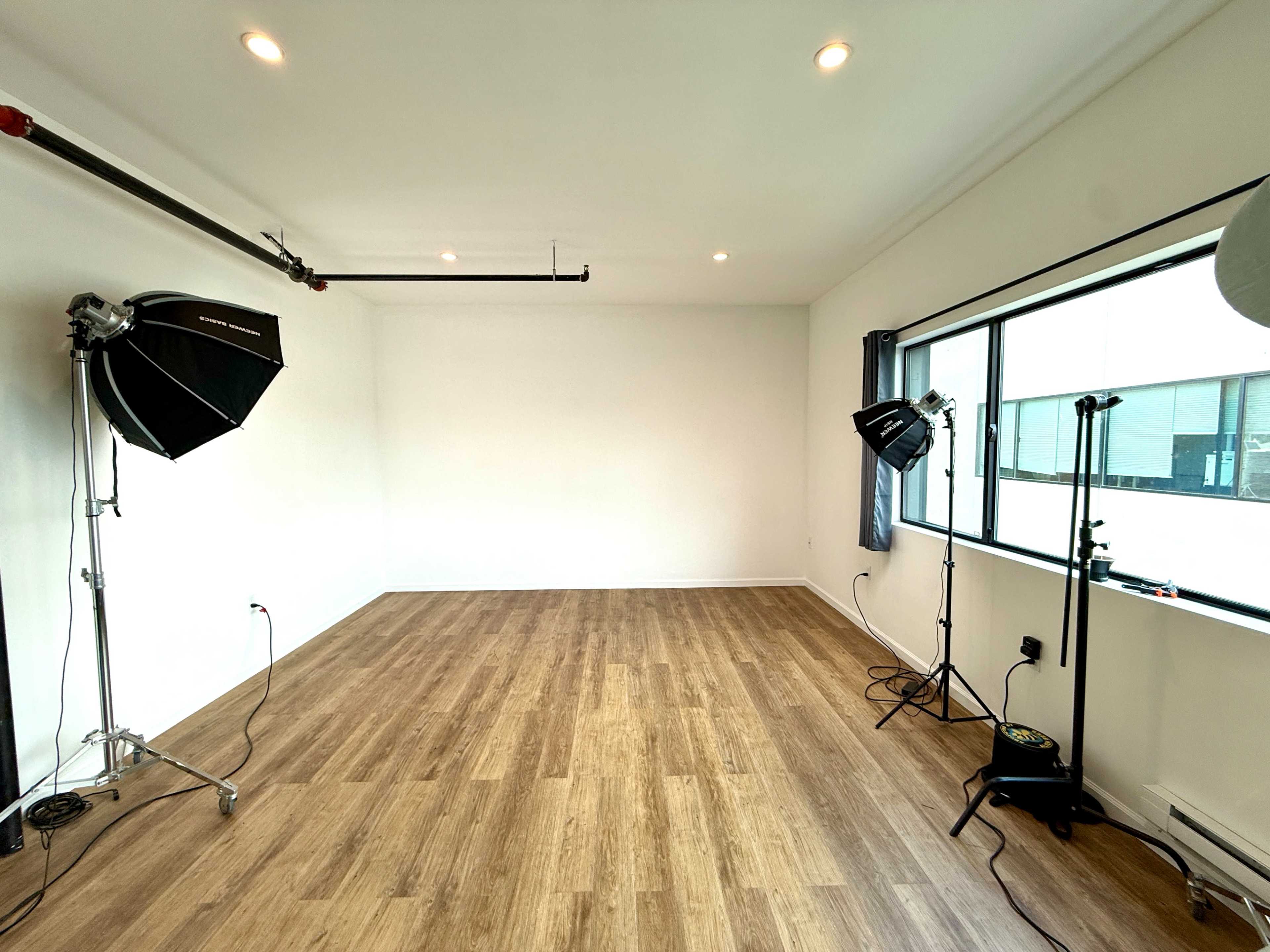 The image shows an empty photo studio with wooden flooring, two light stands with softboxes, and large windows letting in natural light.