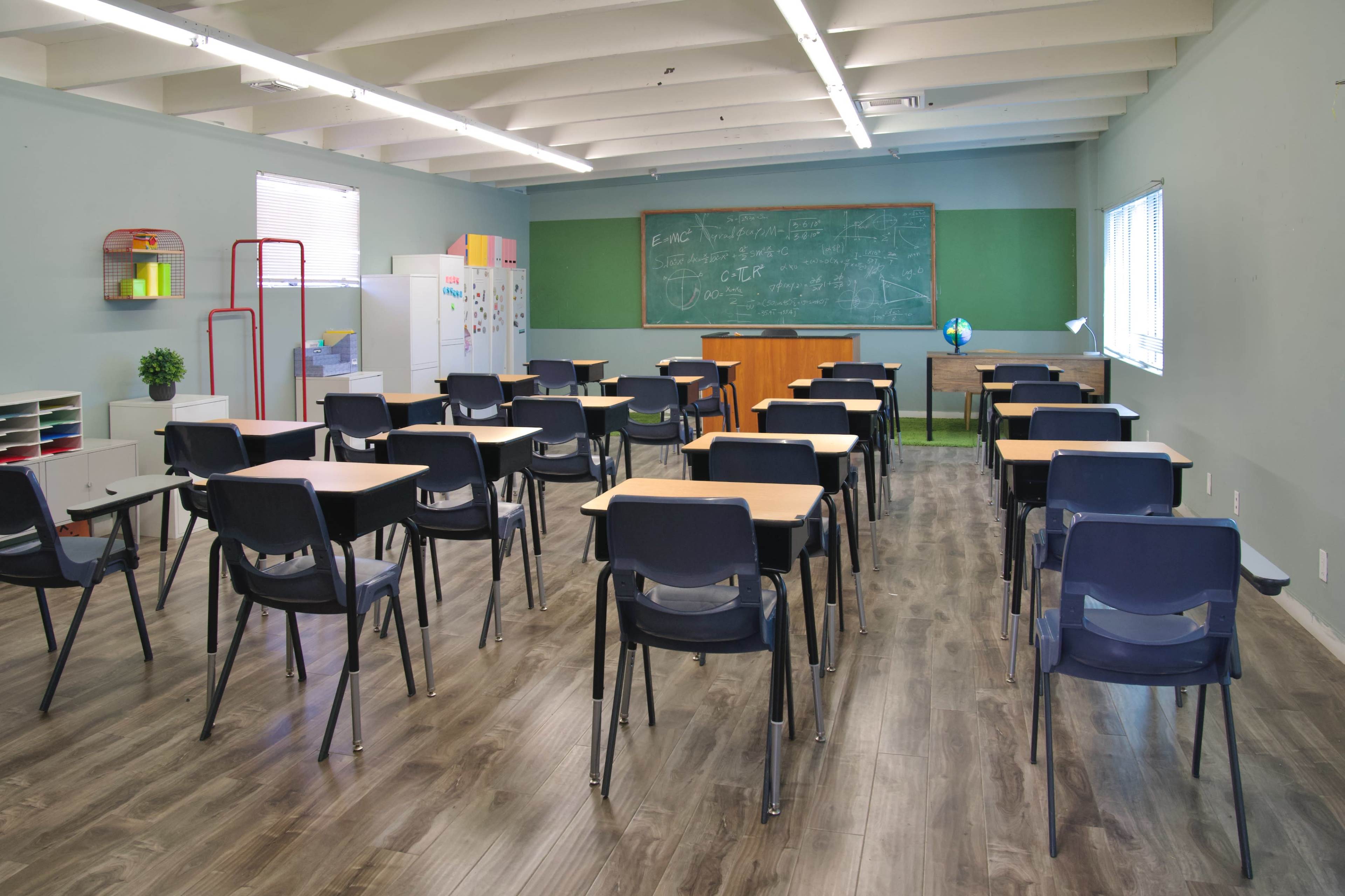 An empty classroom with rows of desks facing a chalkboard and a teacher's desk in front of a green accent wall.