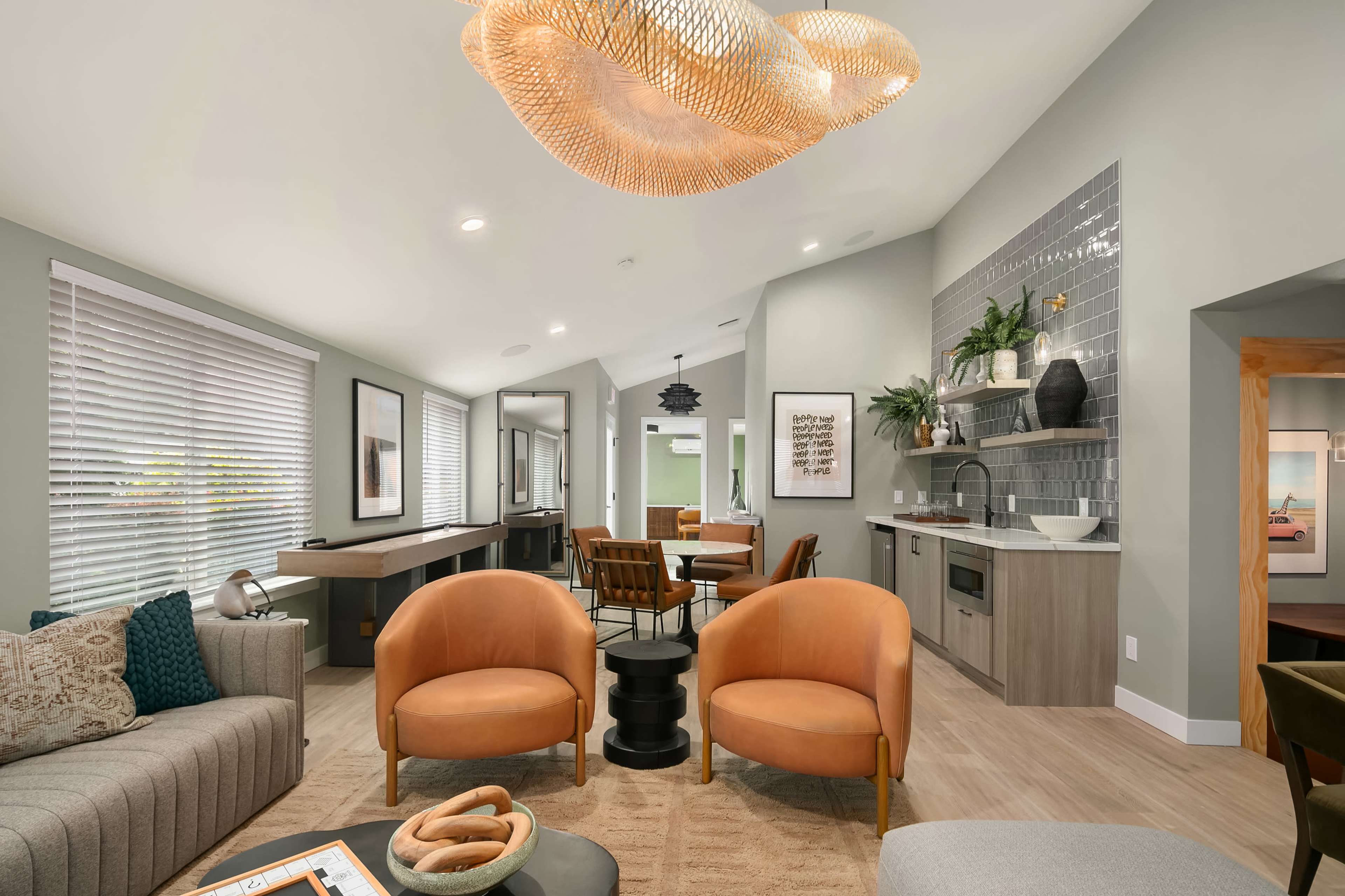 The image shows a modern living space featuring a seating area with orange armchairs, a gray sofa, and a dining area with a round table, all illuminated by a decorative light fixture.