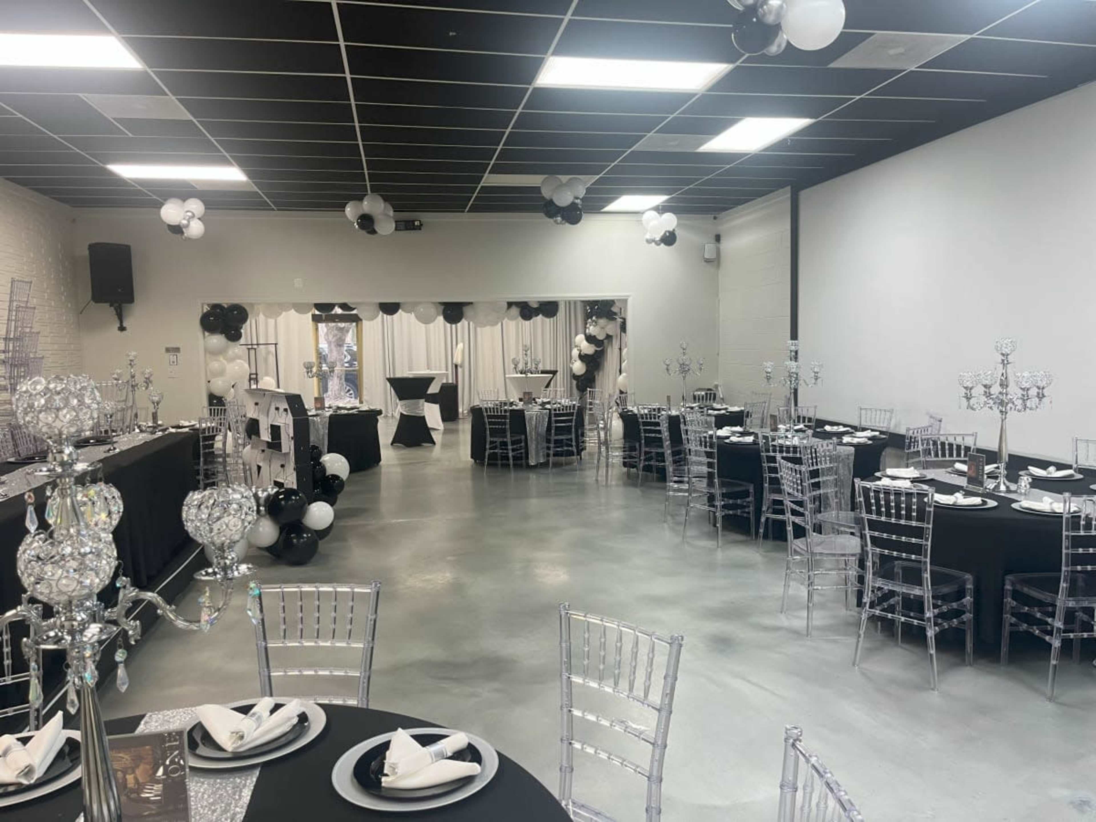 A neat banquet hall is set up for an event, featuring tables dressed in black cloth and silver accents, with decorative centerpieces and balloon arrangements.