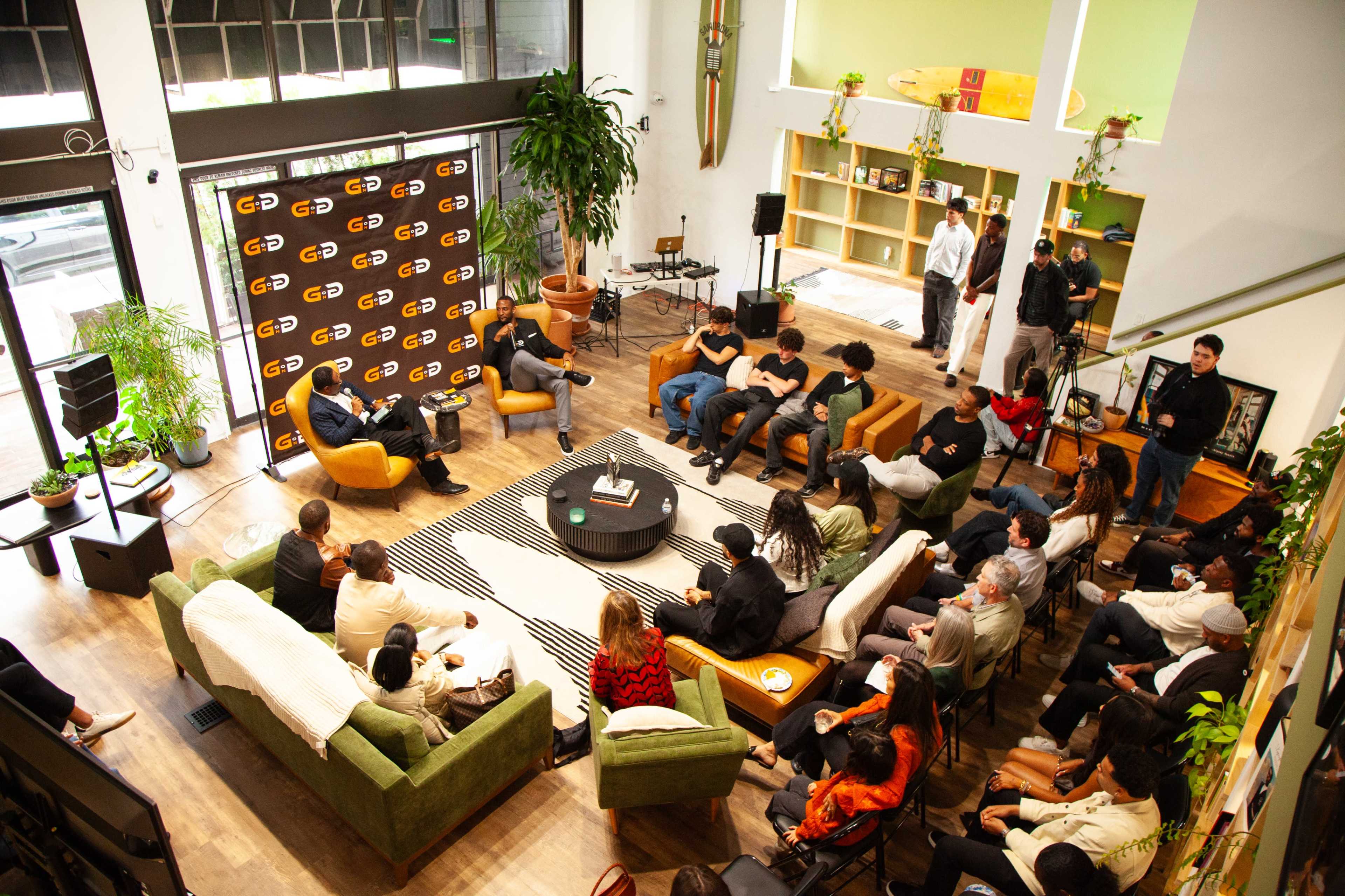 A group of people is seated in a spacious room with large windows, engaging in a discussion while surrounded by plants and modern furniture.