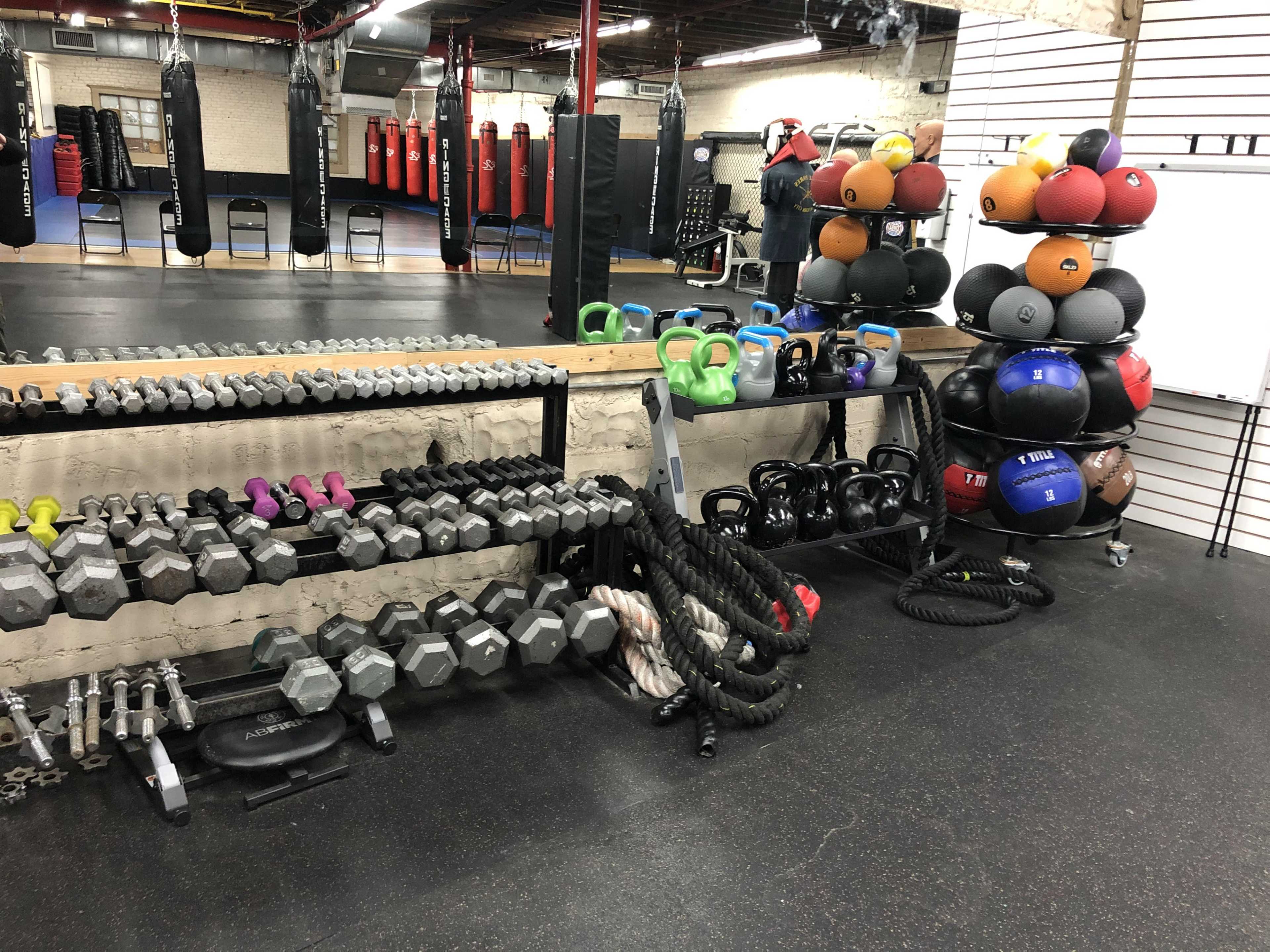 The image shows a fitness area with various weights, kettlebells, medicine balls, and battle ropes arranged neatly along the walls.