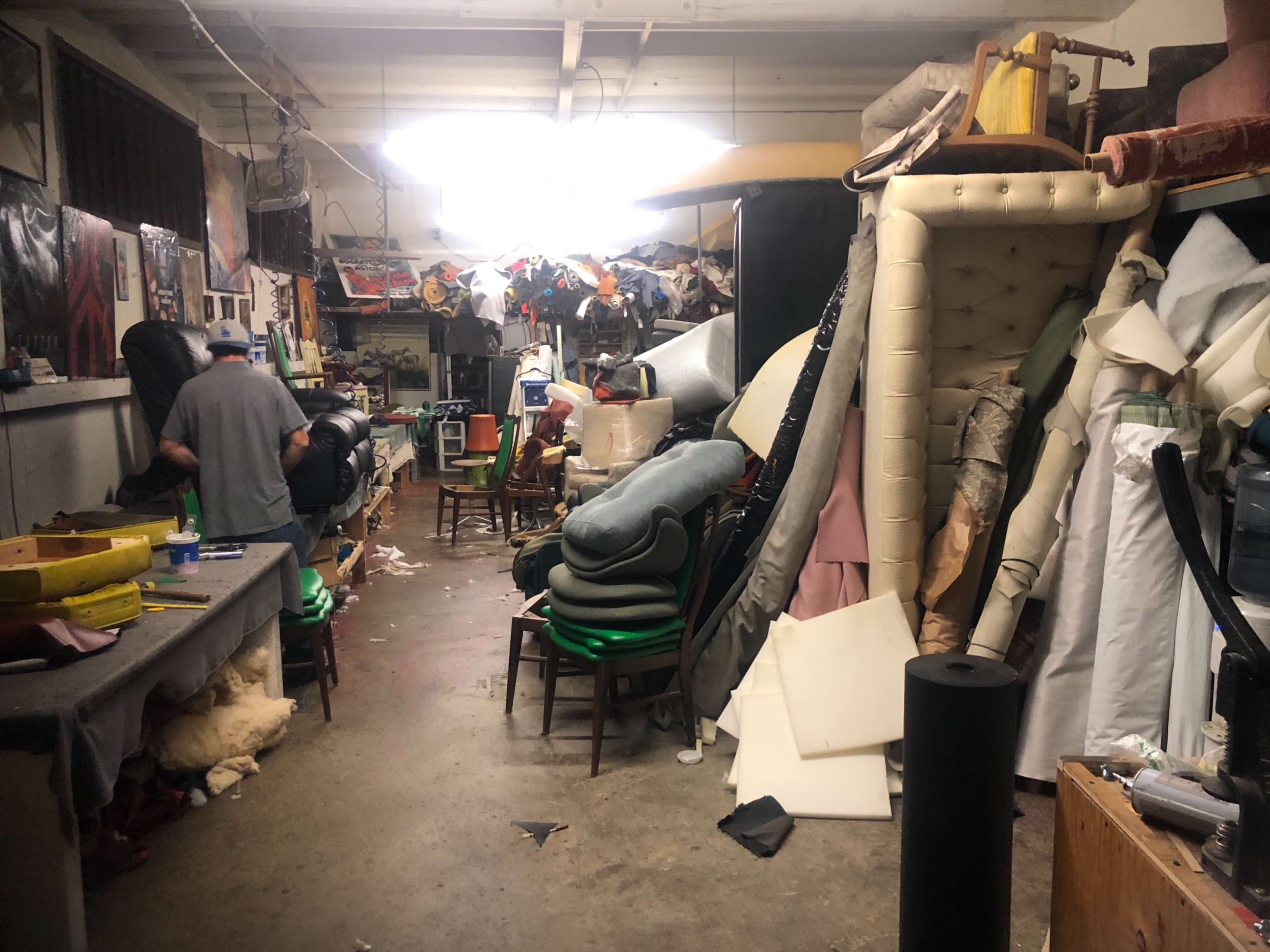 A cluttered workshop is filled with stacked furniture and materials, while a person works in the background.