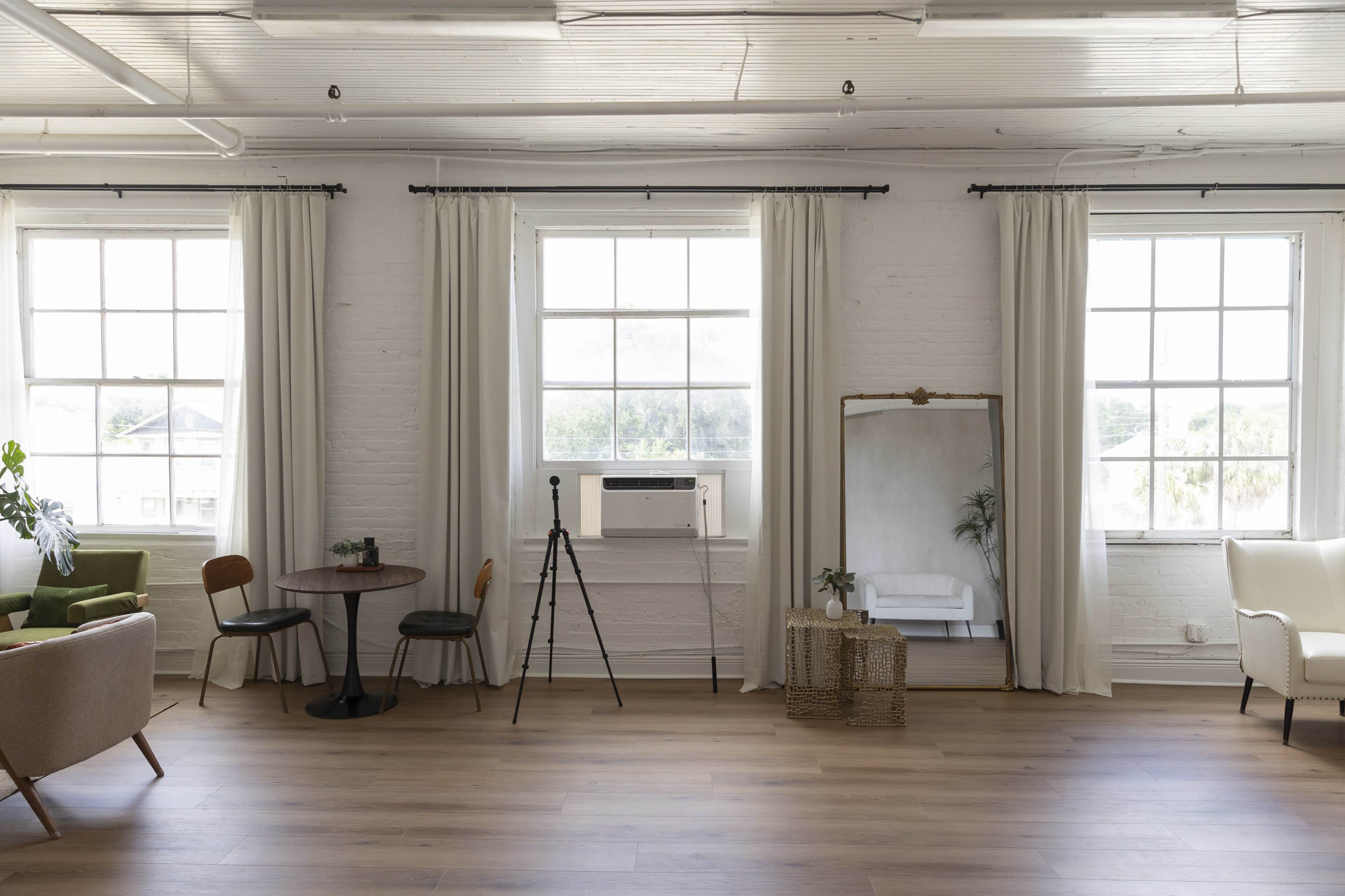 The image shows a bright, spacious room with large windows, light-colored curtains, a mirror, a chair, a small table, and a tripod setup.