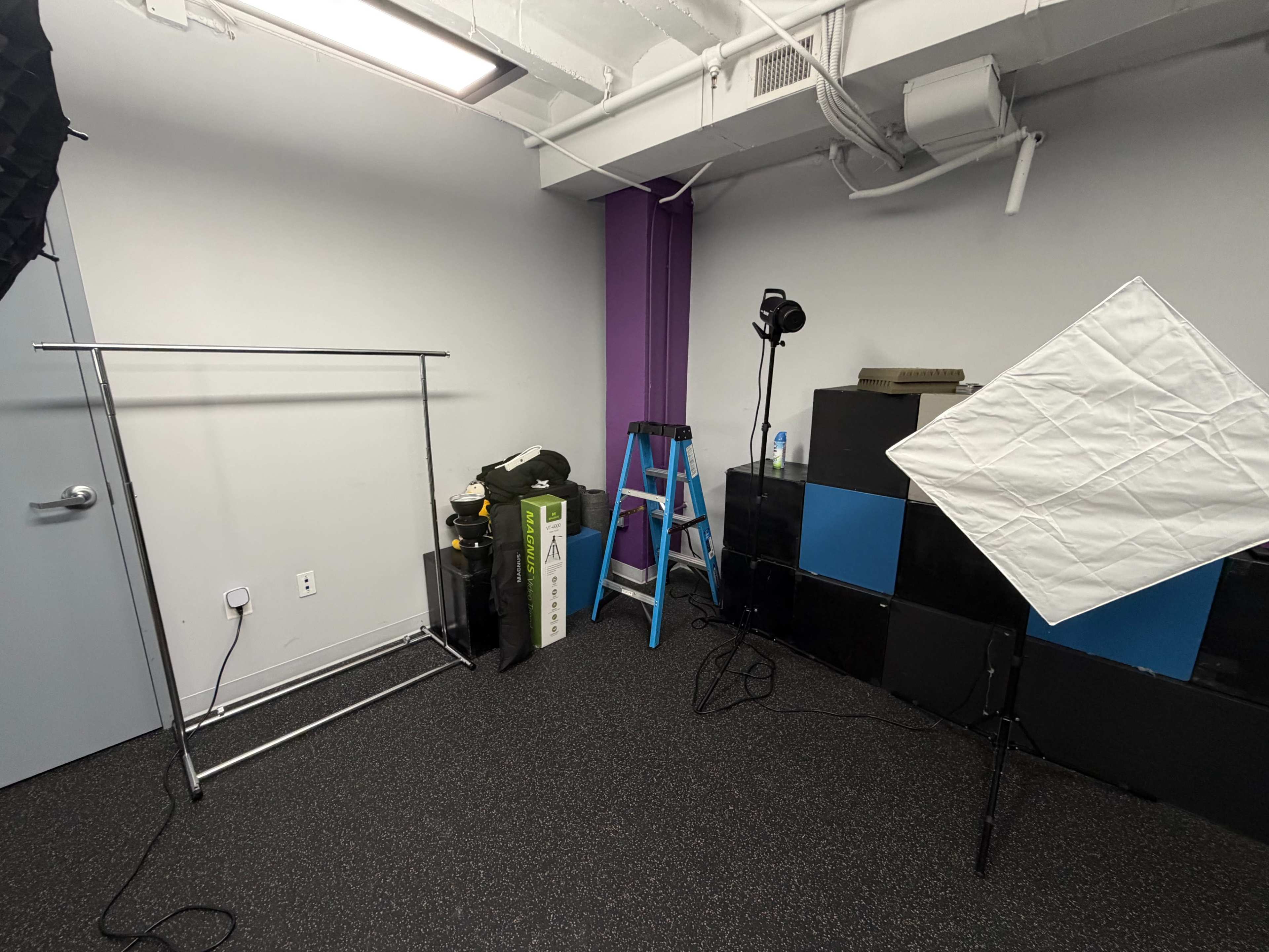 The image shows a small studio space with a clothing rack, a ladder, a light stand, and several storage boxes.