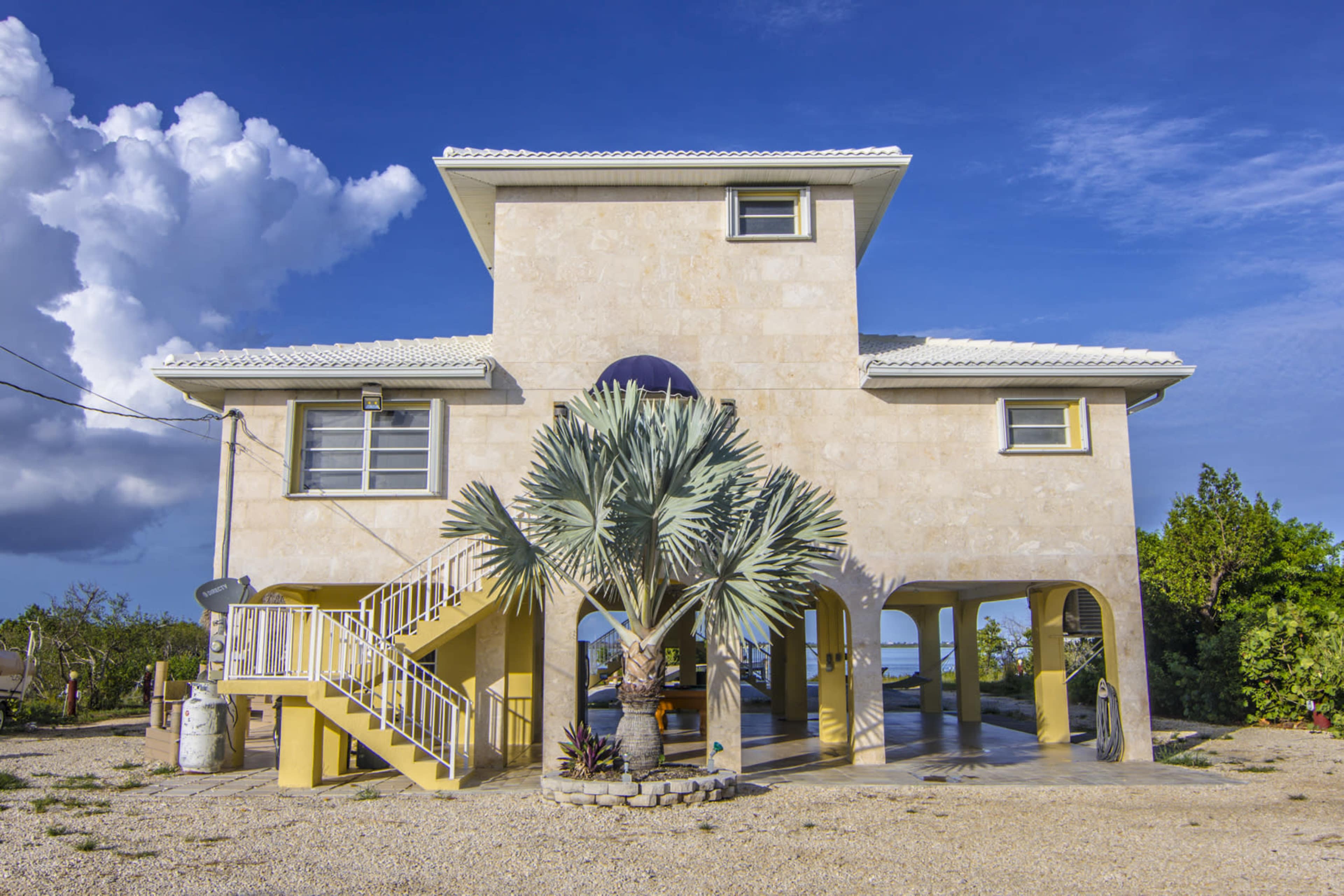 A two-story stone house with a palm tree in front and a staircase leading to the upper floor is set against a blue sky with clouds.