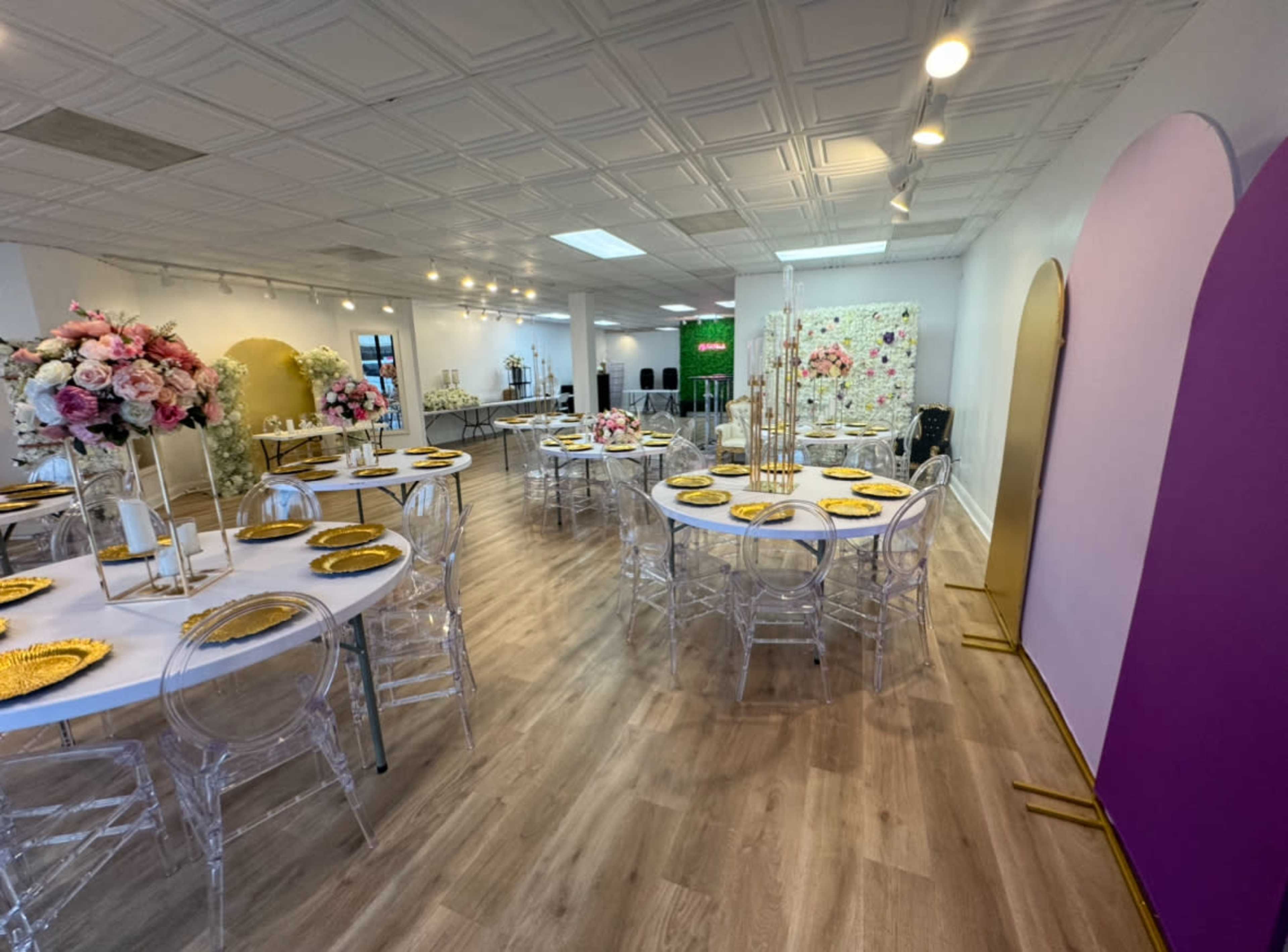 The interior of a spacious event venue features circular tables set with gold plates and transparent chairs, adorned with floral arrangements and illuminated by soft lighting.