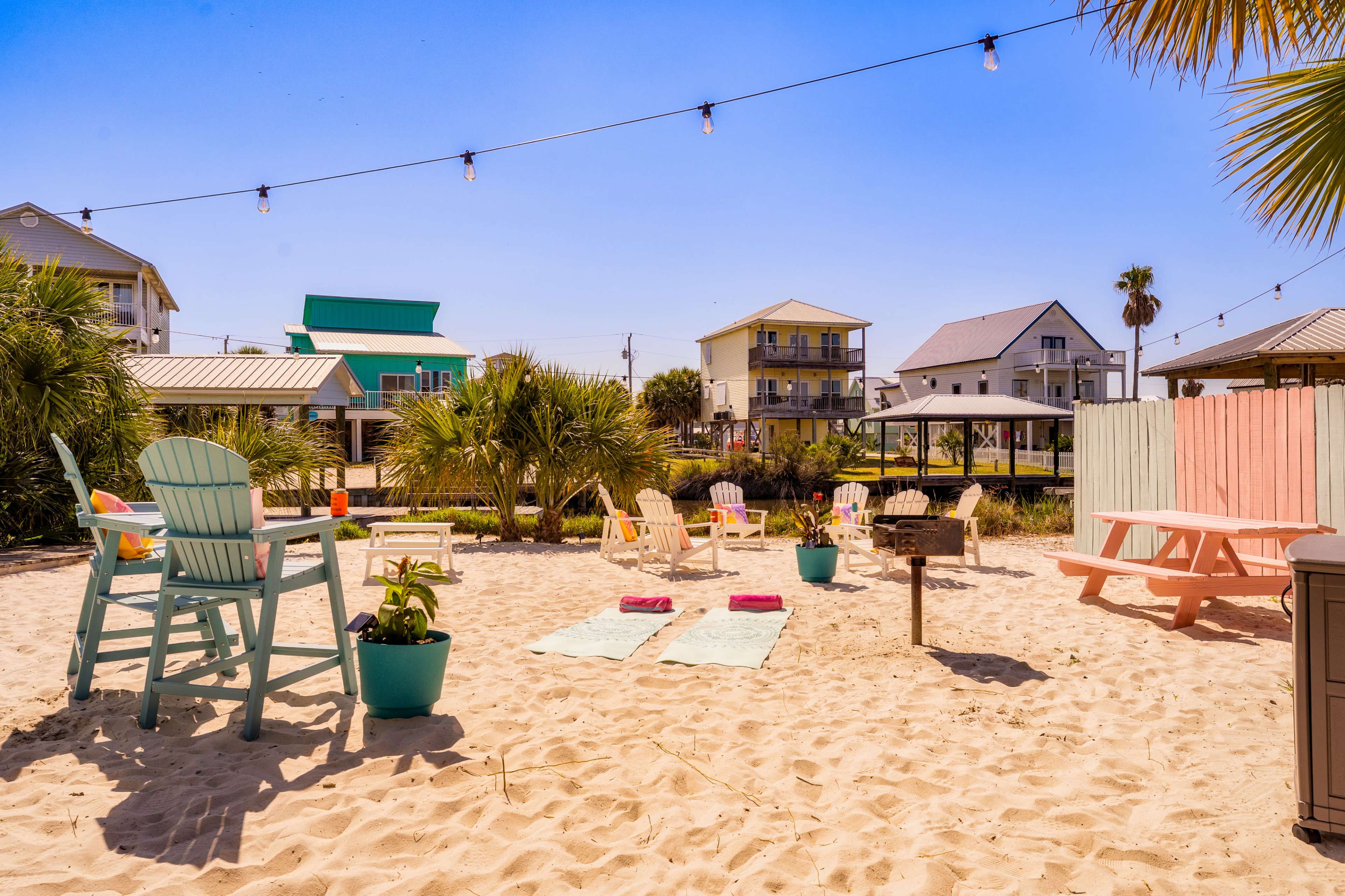 The image shows a sandy outdoor area with lounge chairs, a picnic table, and string lights, surrounded by palm trees and nearby houses.