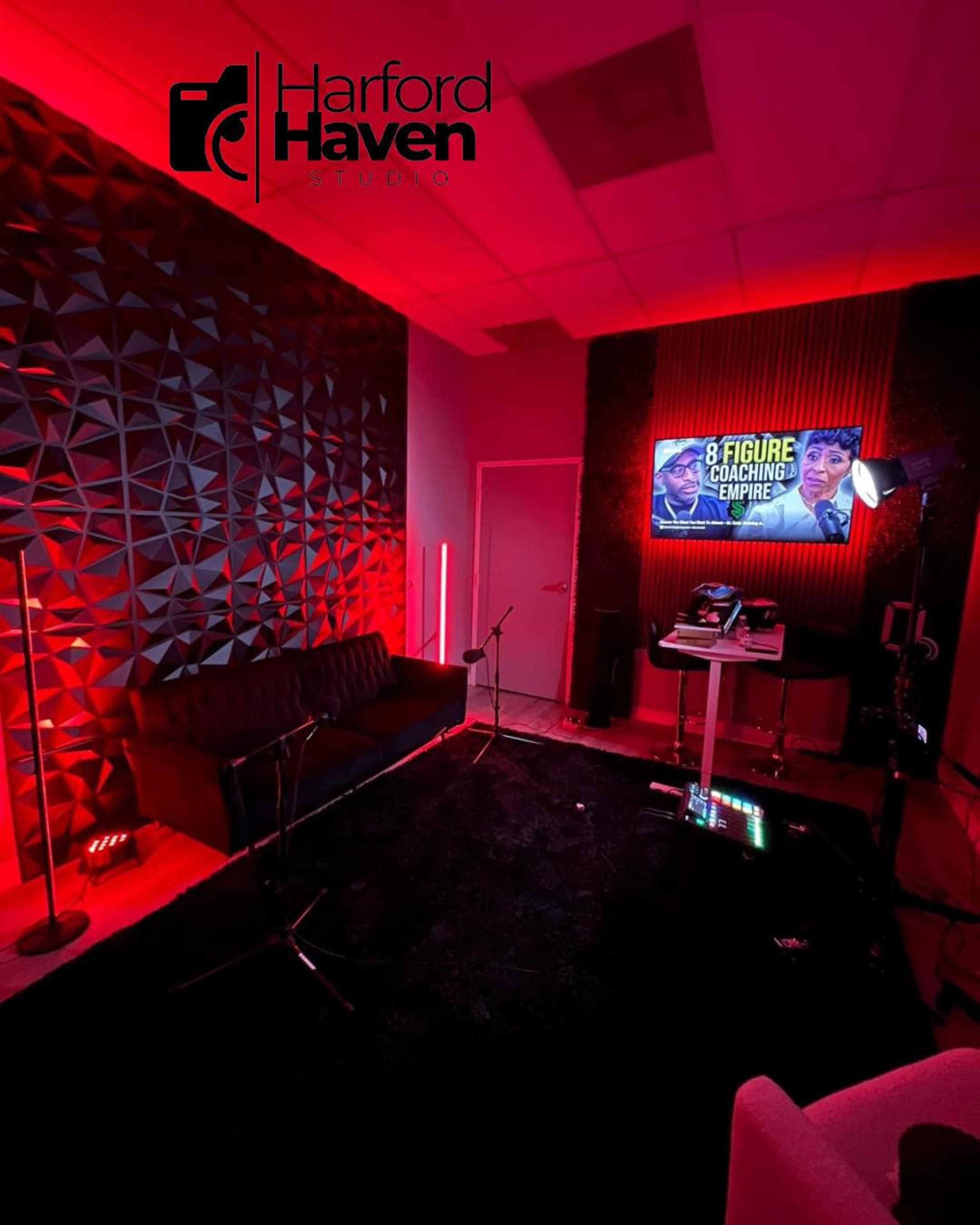 A modern studio space features a dark carpet, a sofa, and a television displaying a coaching program, all illuminated by red lighting.