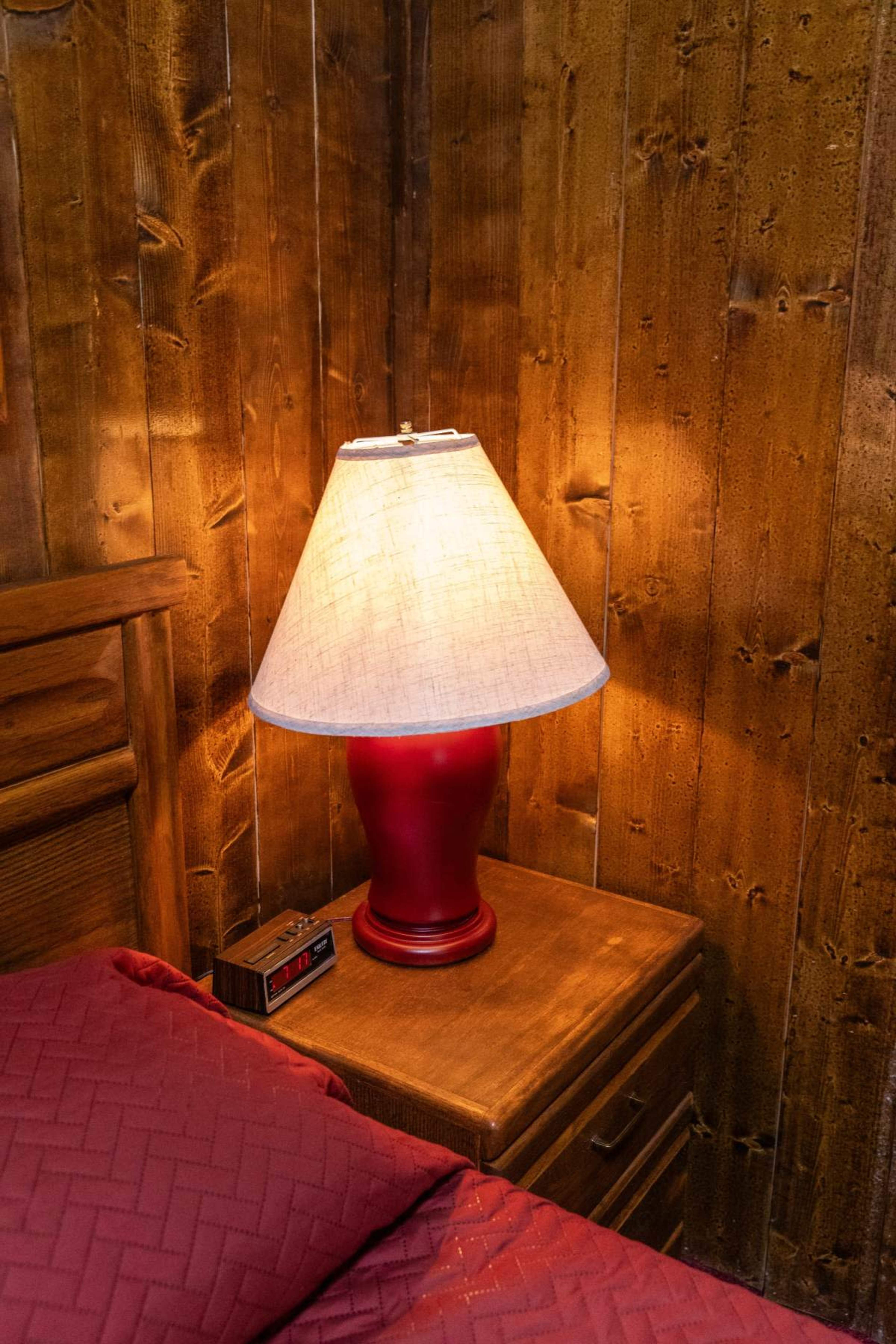 A red lamp with a fabric shade sits on a wooden nightstand beside a bed with a burgundy coverlet, illuminated against a backdrop of wooden walls.