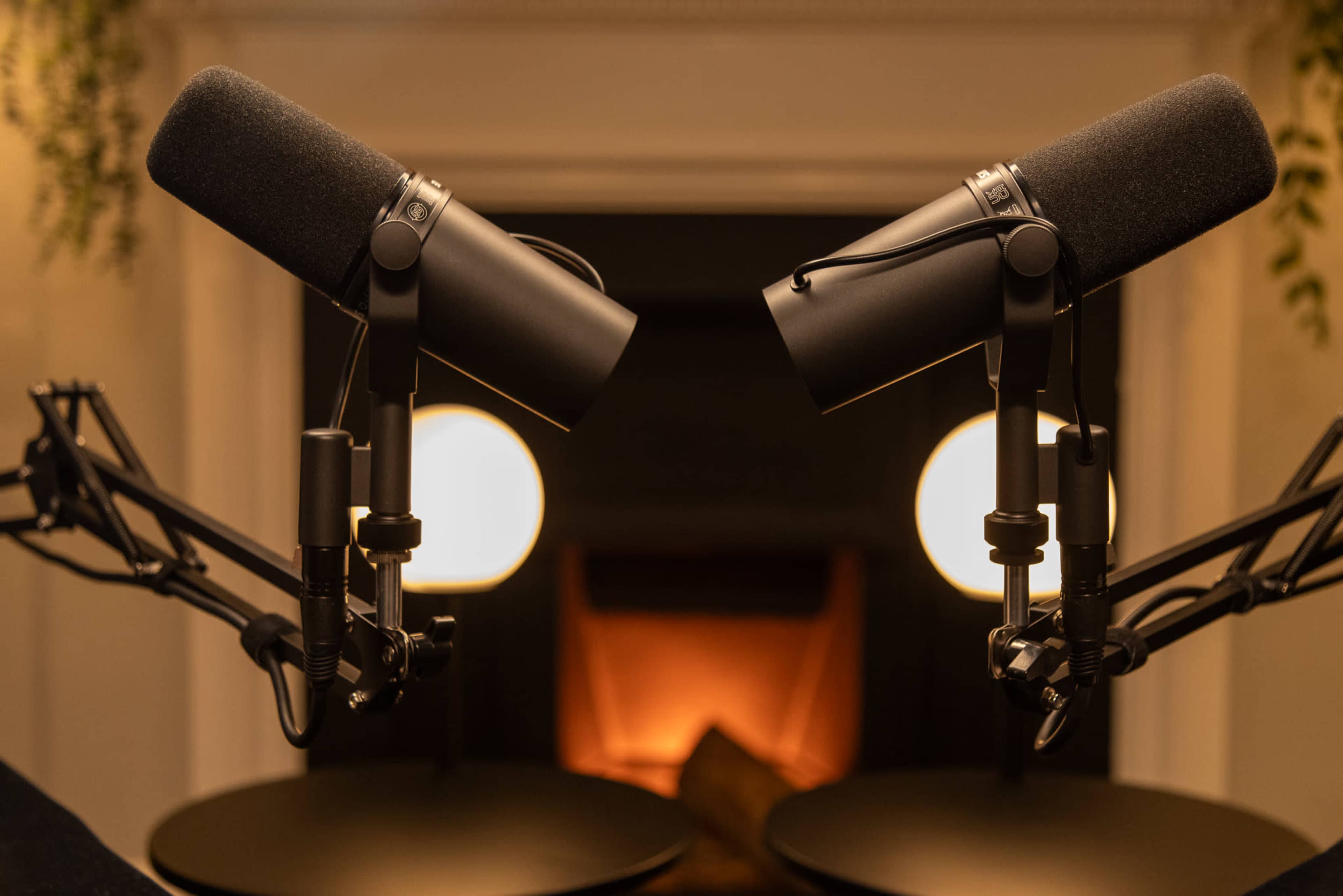 Two microphones are positioned on stands facing each other, with a softly lit fireplace in the background.