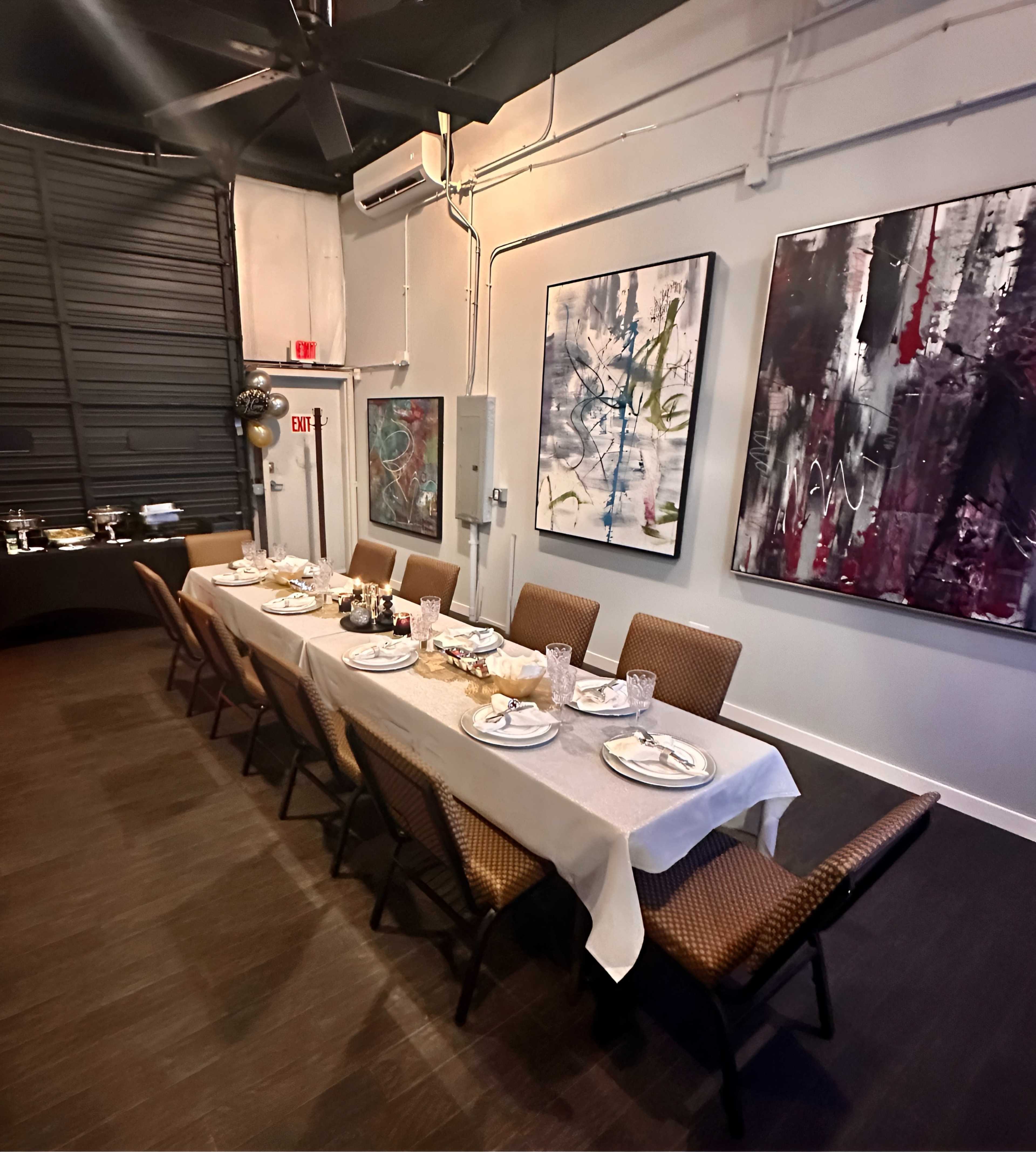 A long dining table is set with plates, utensils, and decorative elements, positioned in a modern room featuring abstract artwork on the walls.