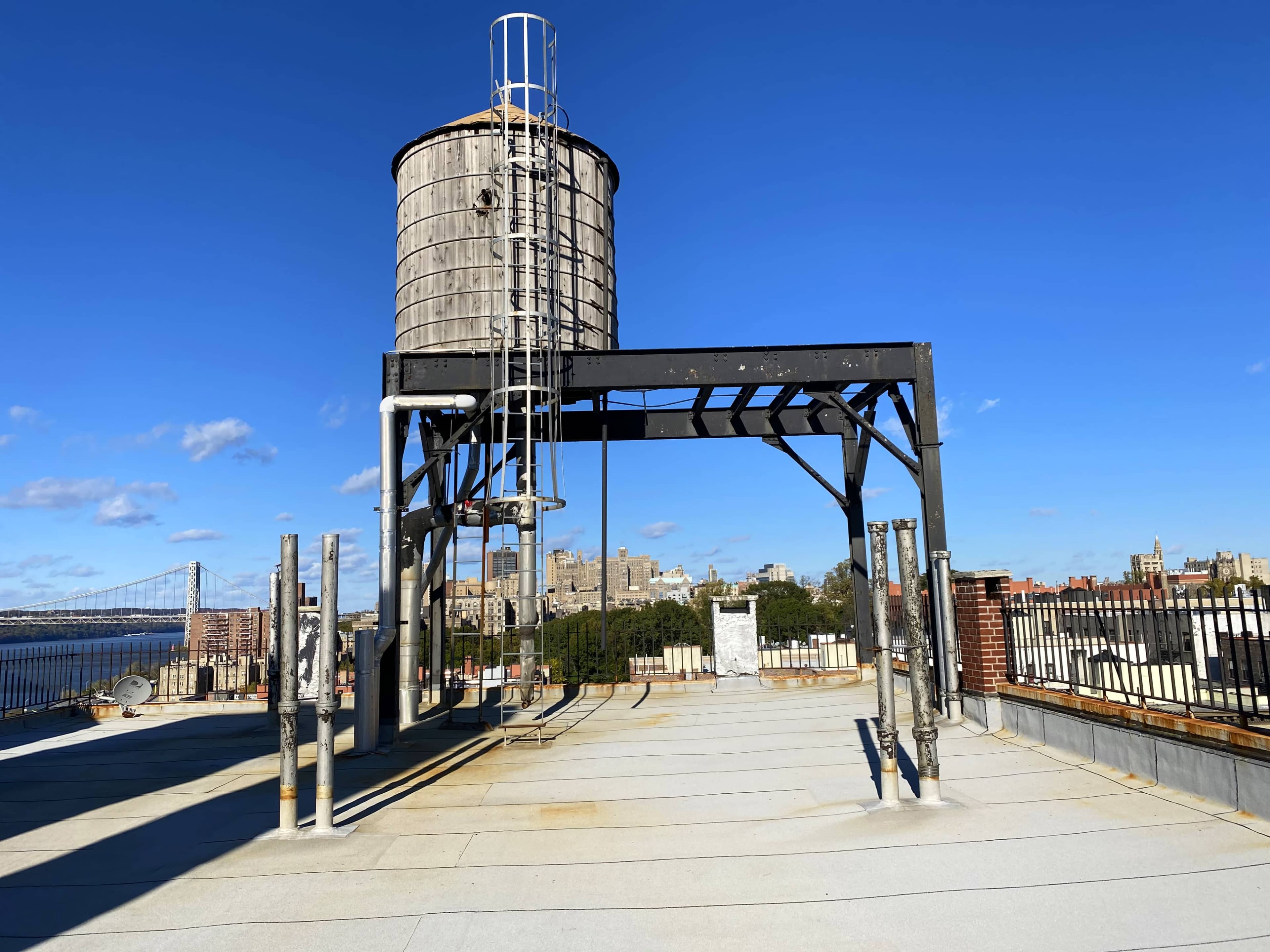 A wooden water tower stands on a rooftop with a clear blue sky and a city skyline in the background.