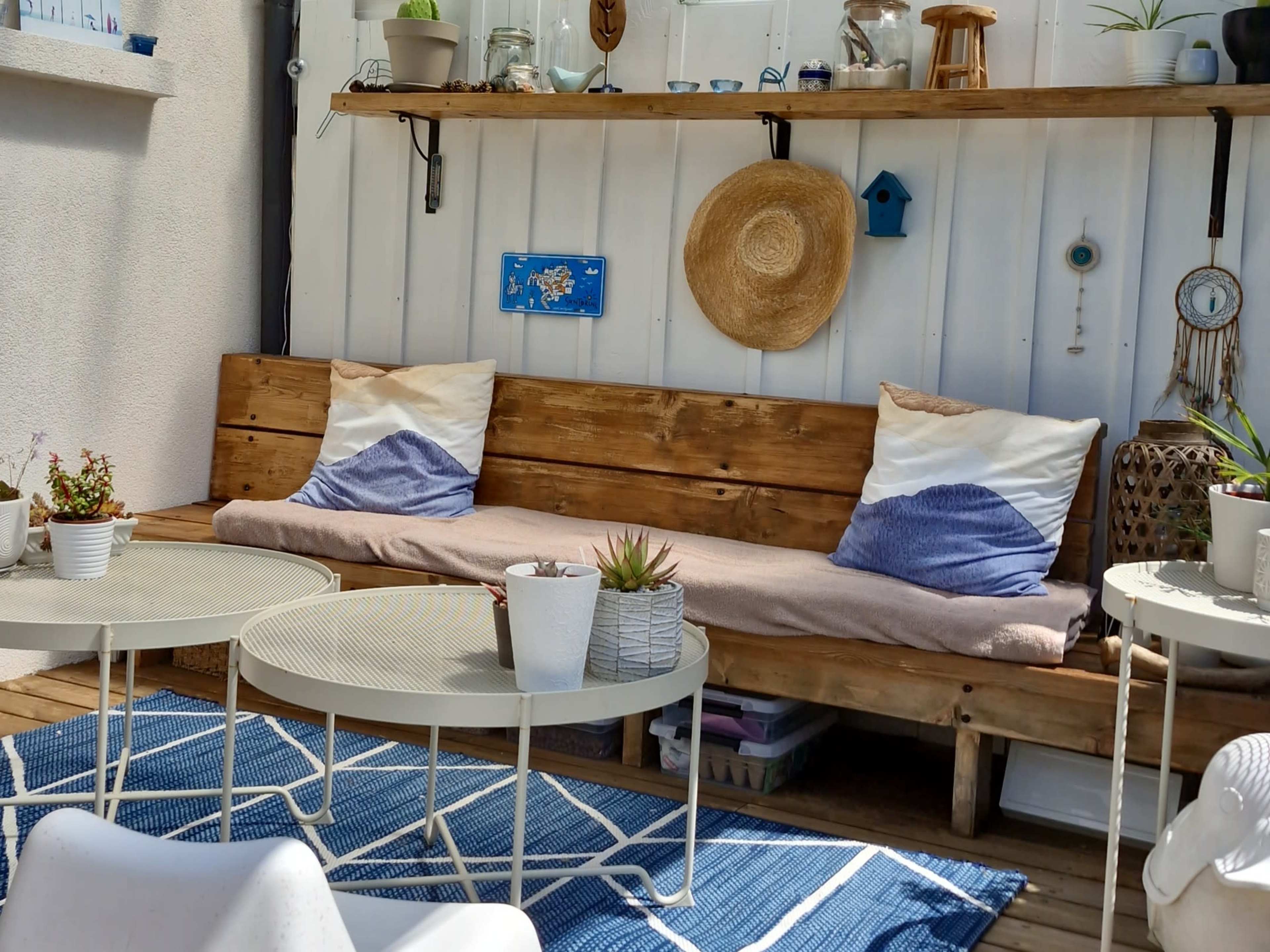 The image shows a cozy outdoor seating area with a wooden bench decorated with cushions, a shelf holding various decorative items, and a round table on a blue rug.