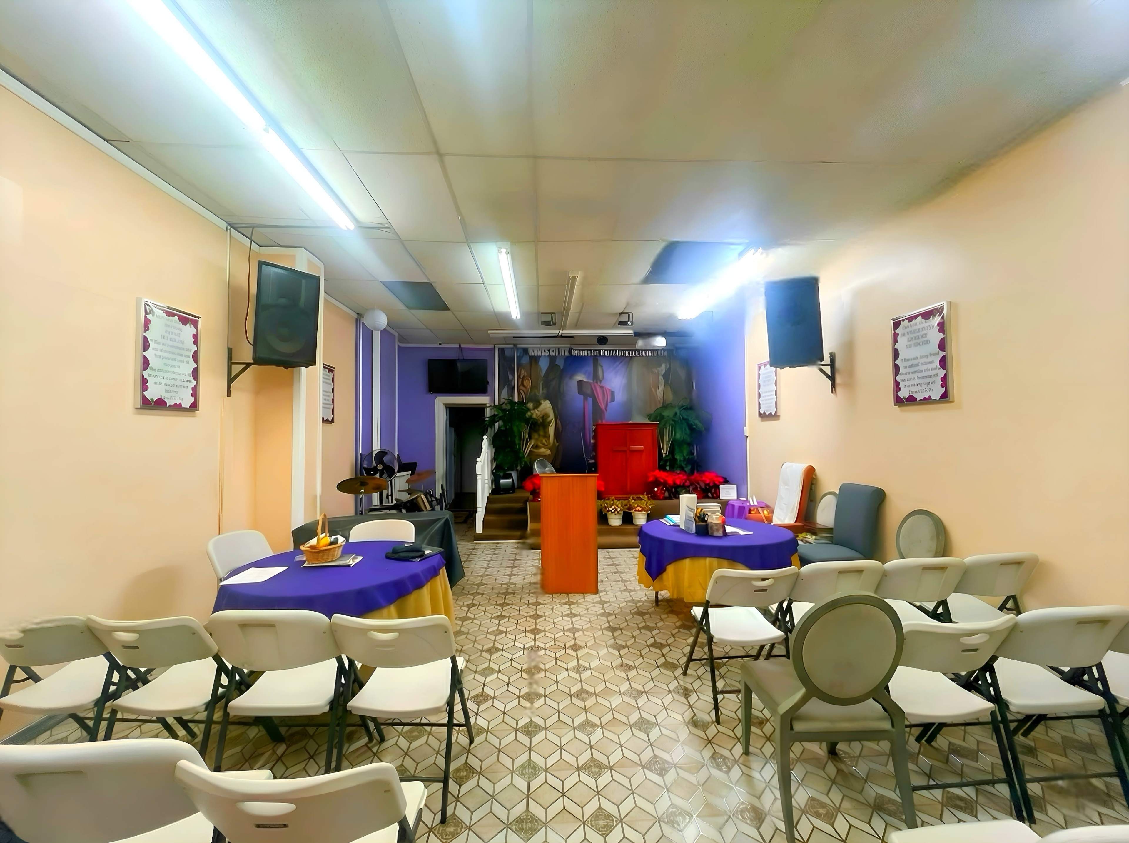 The image shows a small indoor space set up for a gathering, featuring rows of folding chairs, tables covered with cloth, and a backdrop with religious imagery.
