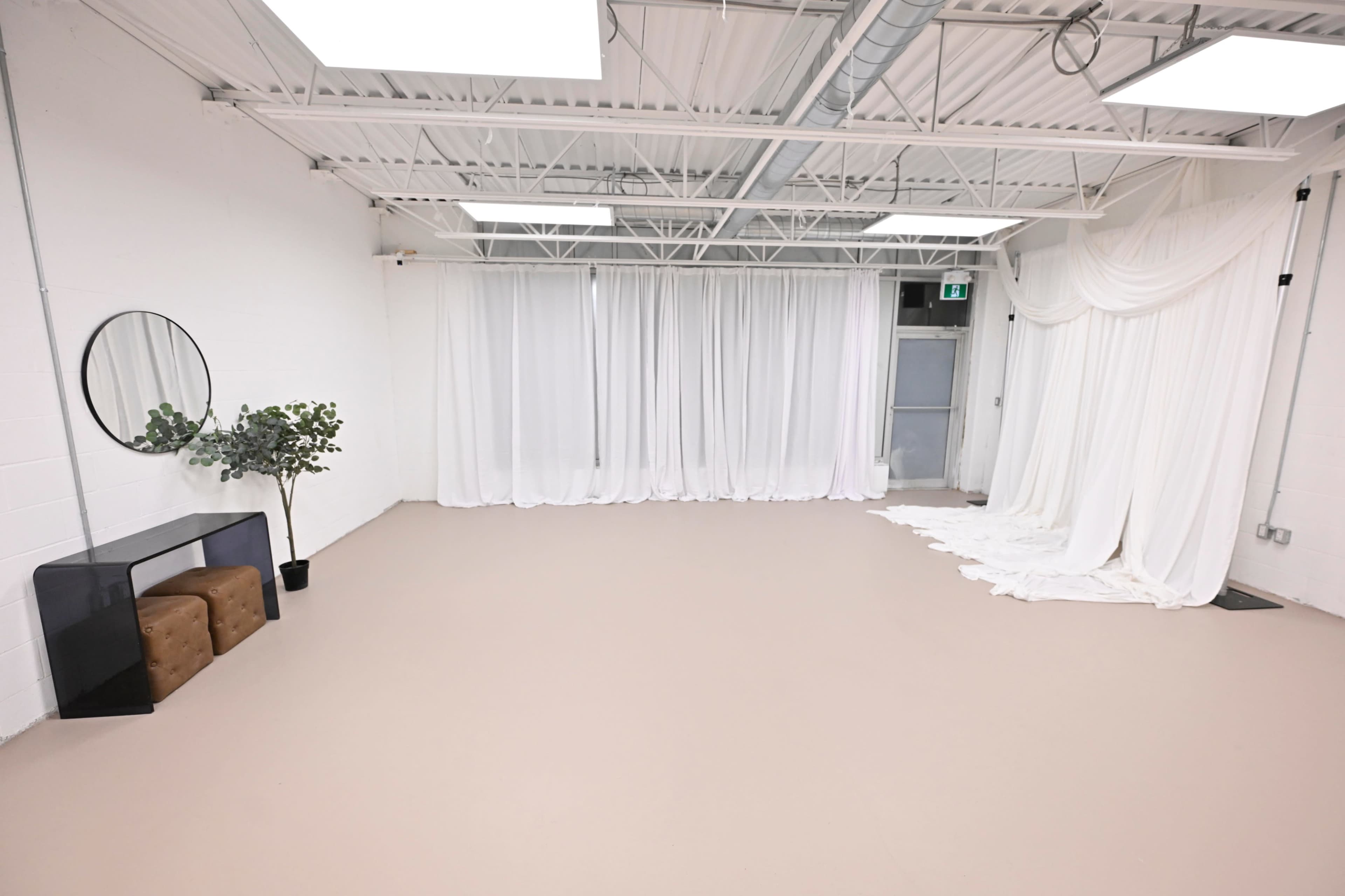 The image shows a spacious, minimalist room with light-colored flooring, white drapes, a mirror, a plant, and a small brown ottoman.