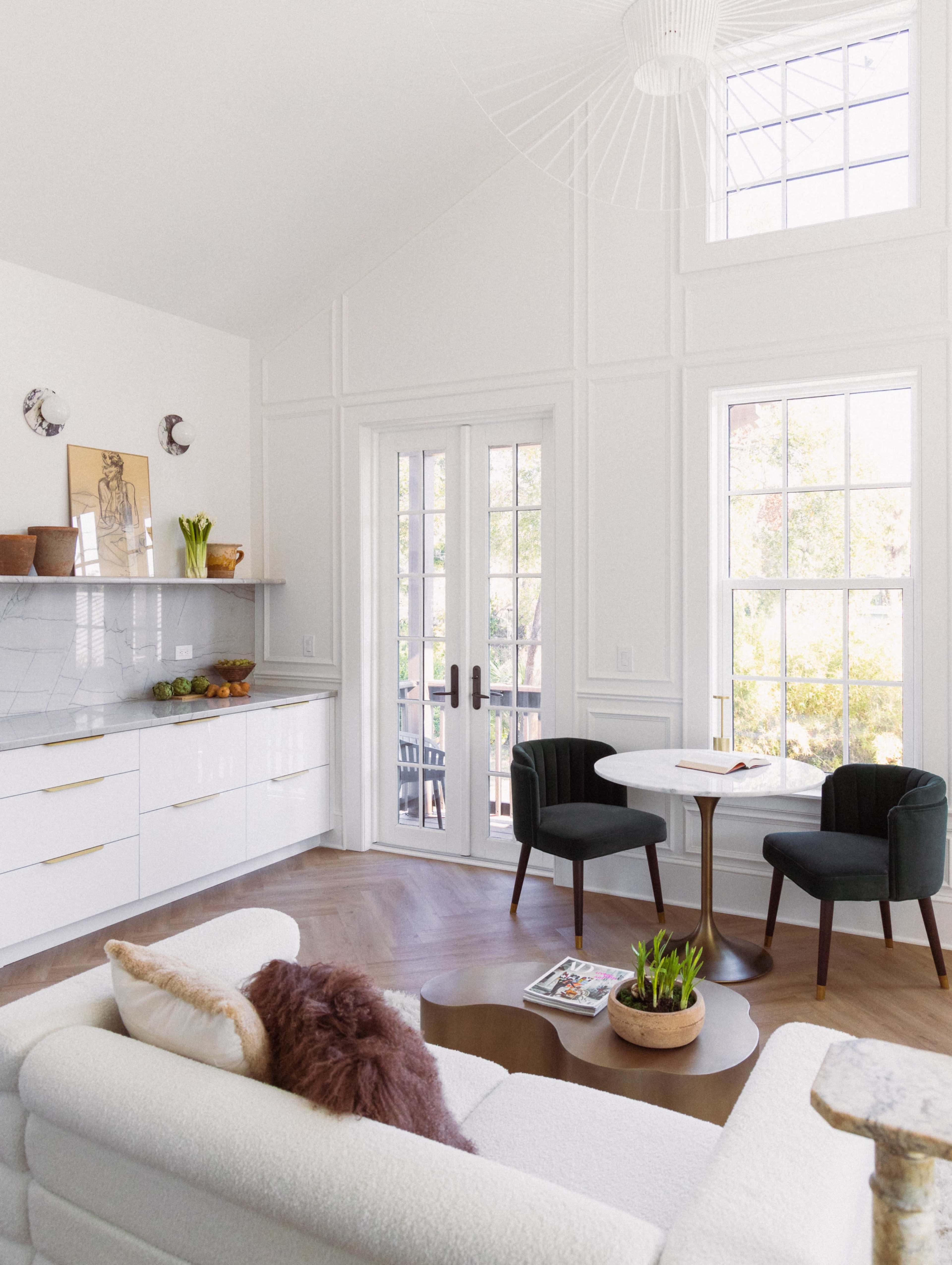 The living room features a light-colored sofa, a round coffee table, and two chairs next to a large window and French doors.