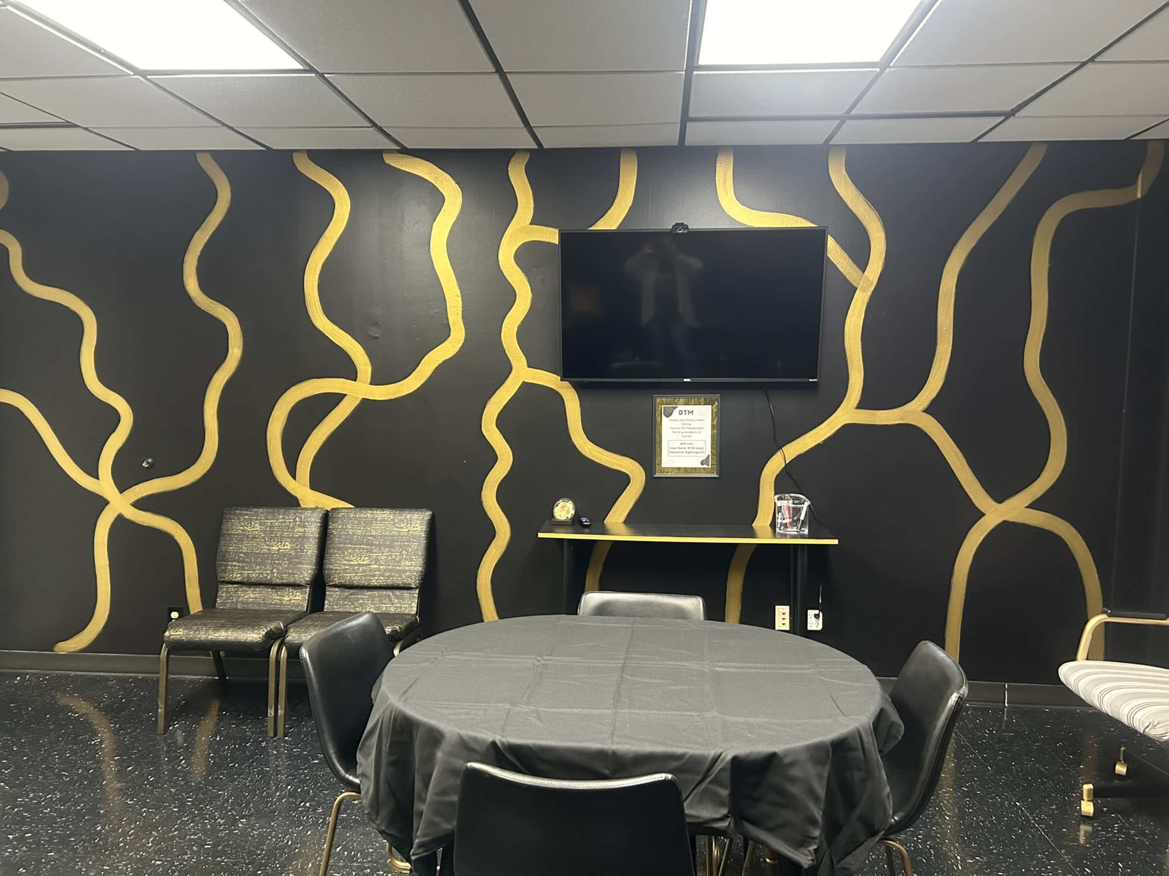 A room with a black wall featuring gold wavy patterns, a television mounted on the wall, and a round black table surrounded by chairs.
