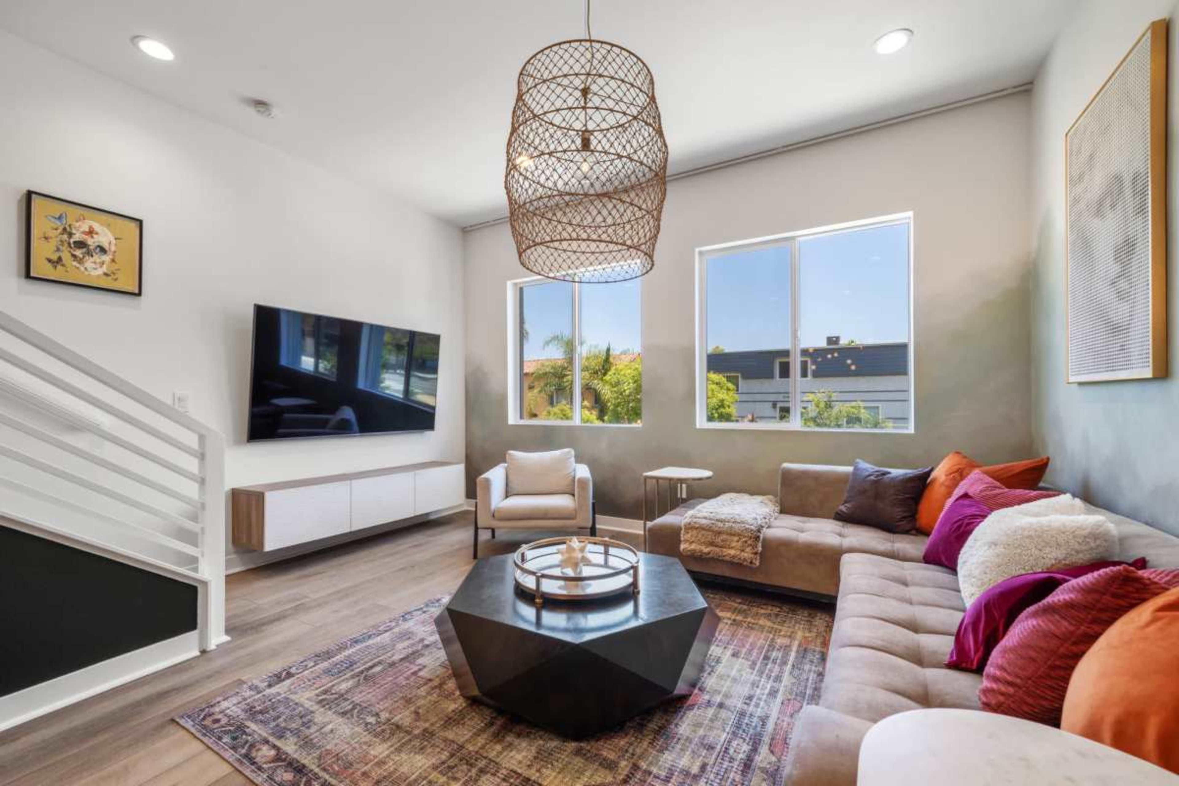 A modern living room features a sectional sofa, a large flat-screen TV on the wall, and a geometric coffee table under a woven pendant chandelier.