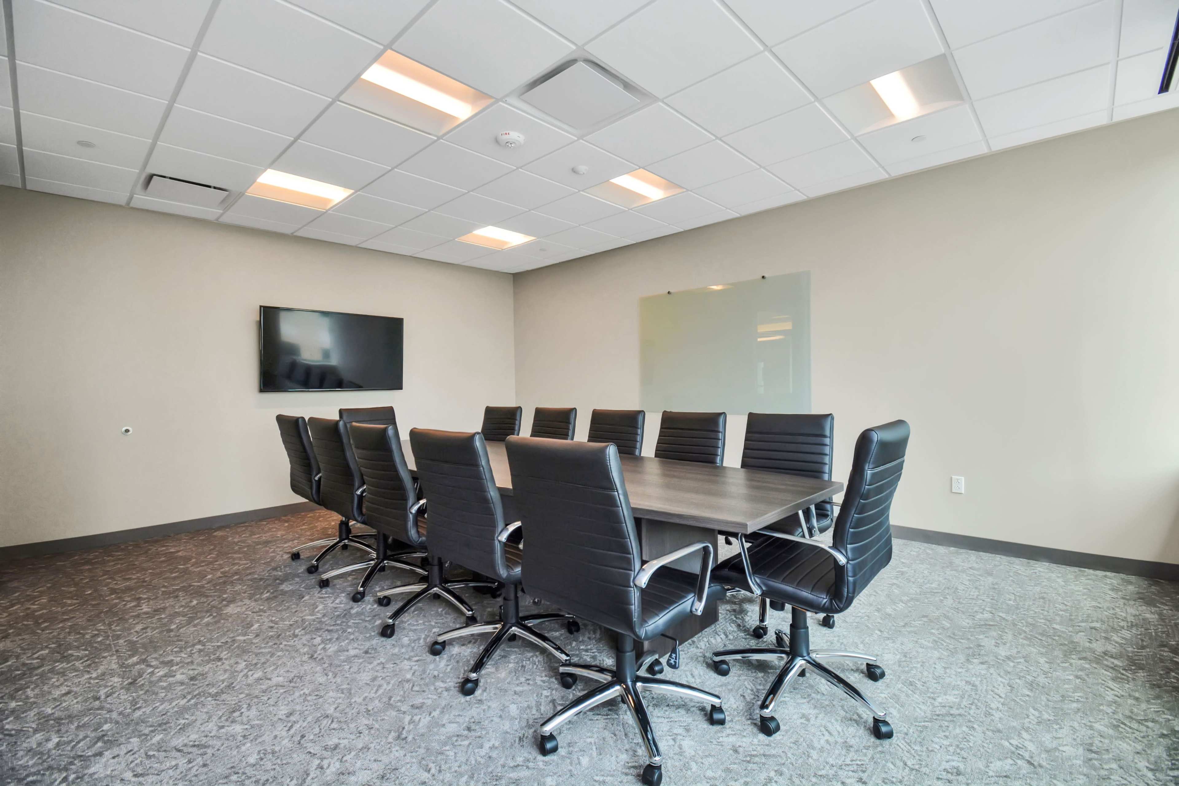 A conference room features a long table surrounded by black leather chairs, with a wall-mounted screen and a glass writing board.