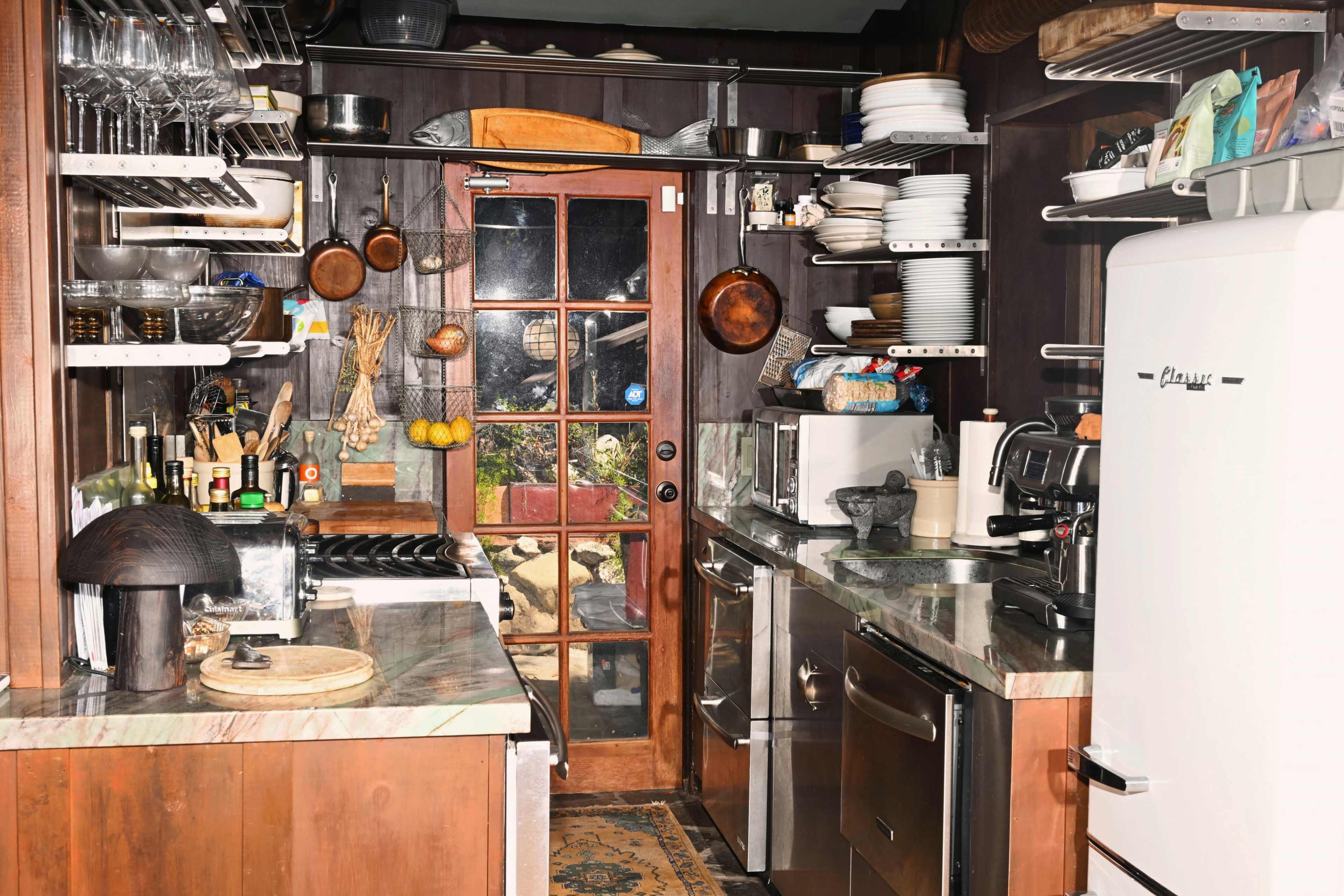 The image shows a compact kitchen with wooden cabinetry, various cooking utensils, a vintage refrigerator, and a glass door leading to an outdoor area.