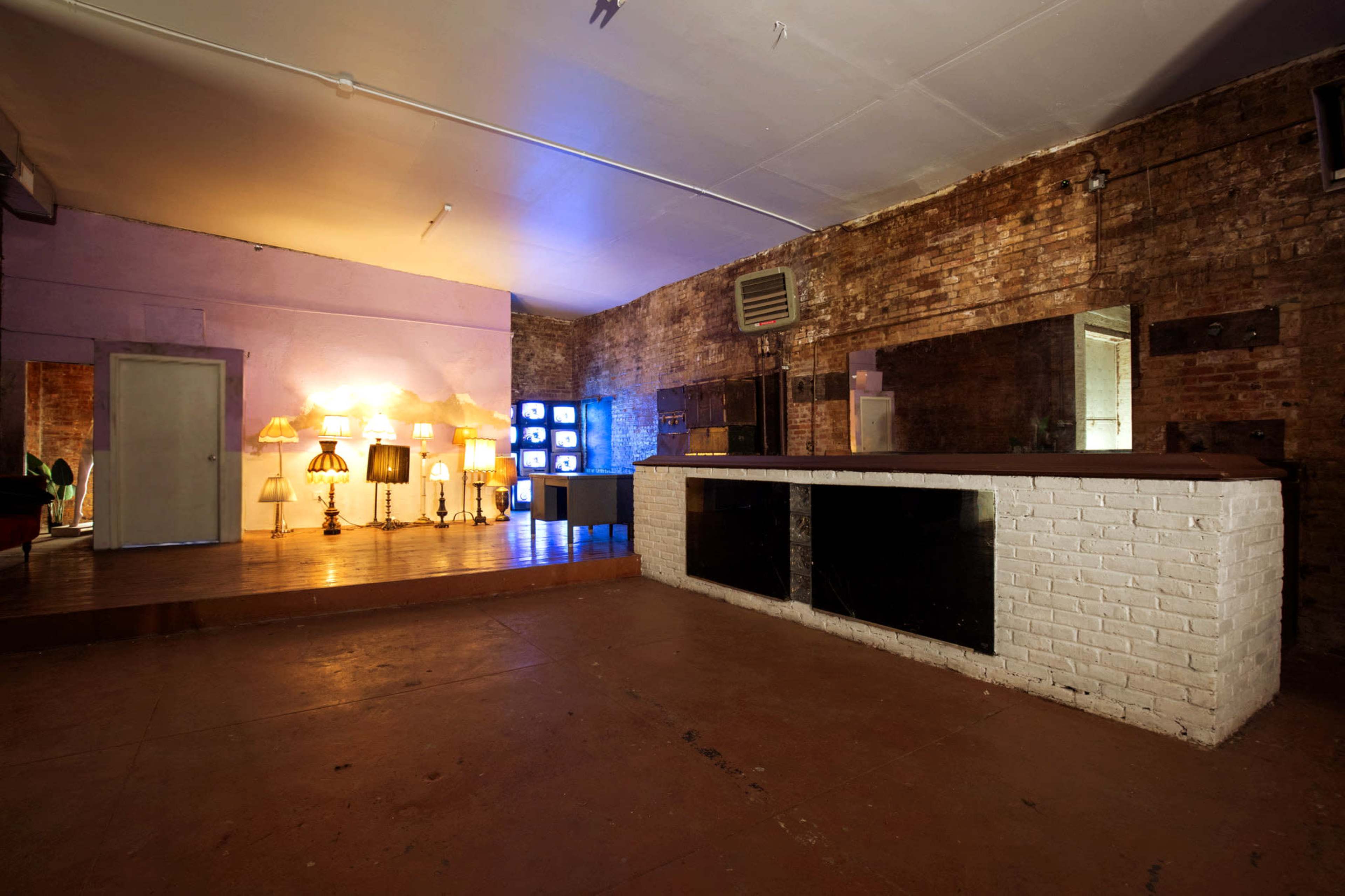 The image shows an empty, industrial-style room with exposed brick walls, a bar area, and several decorative lamps along one wall.