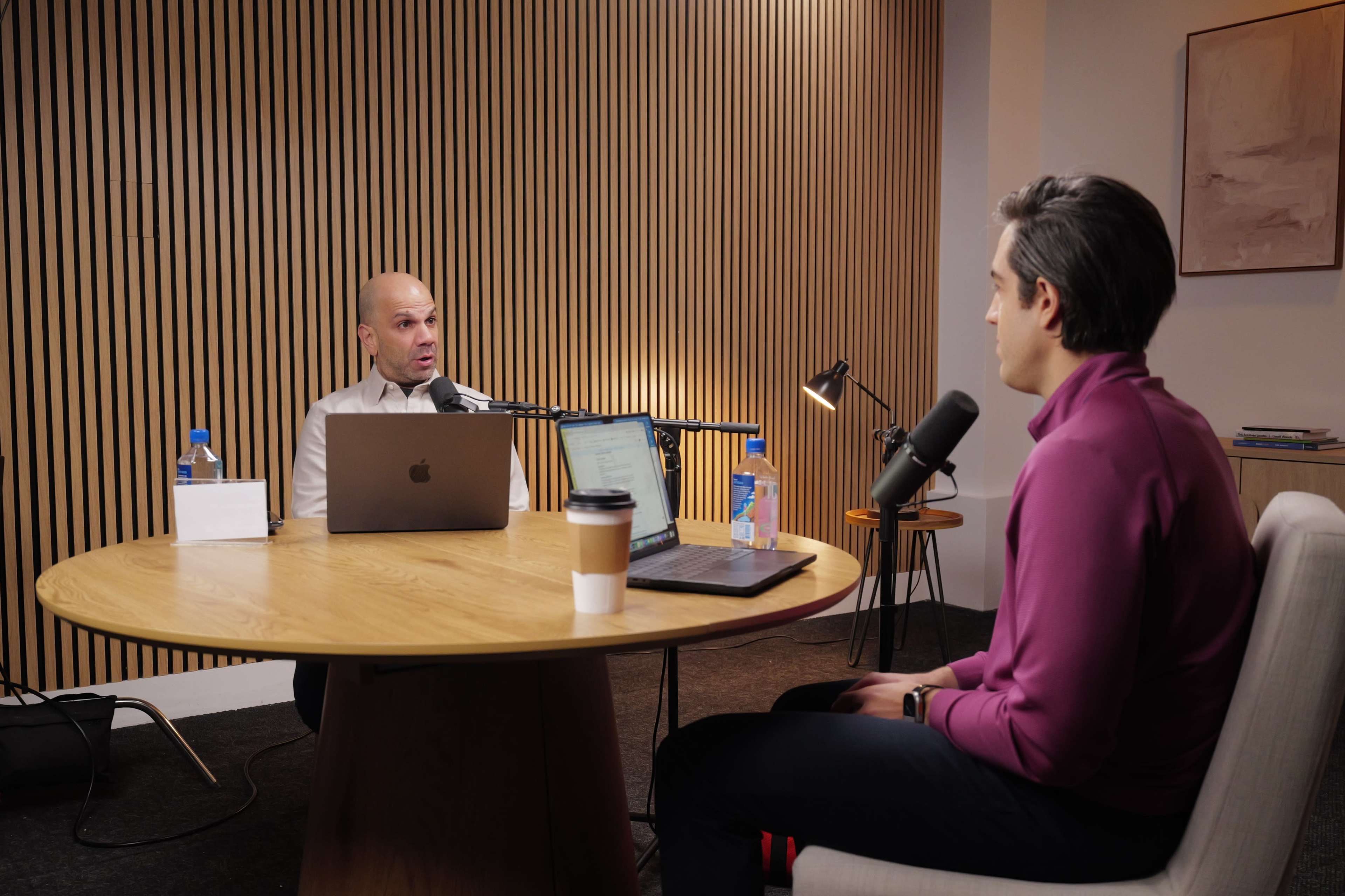 Two men sit at a round wooden table in a modern room, engaging in conversation while using laptops and microphones.