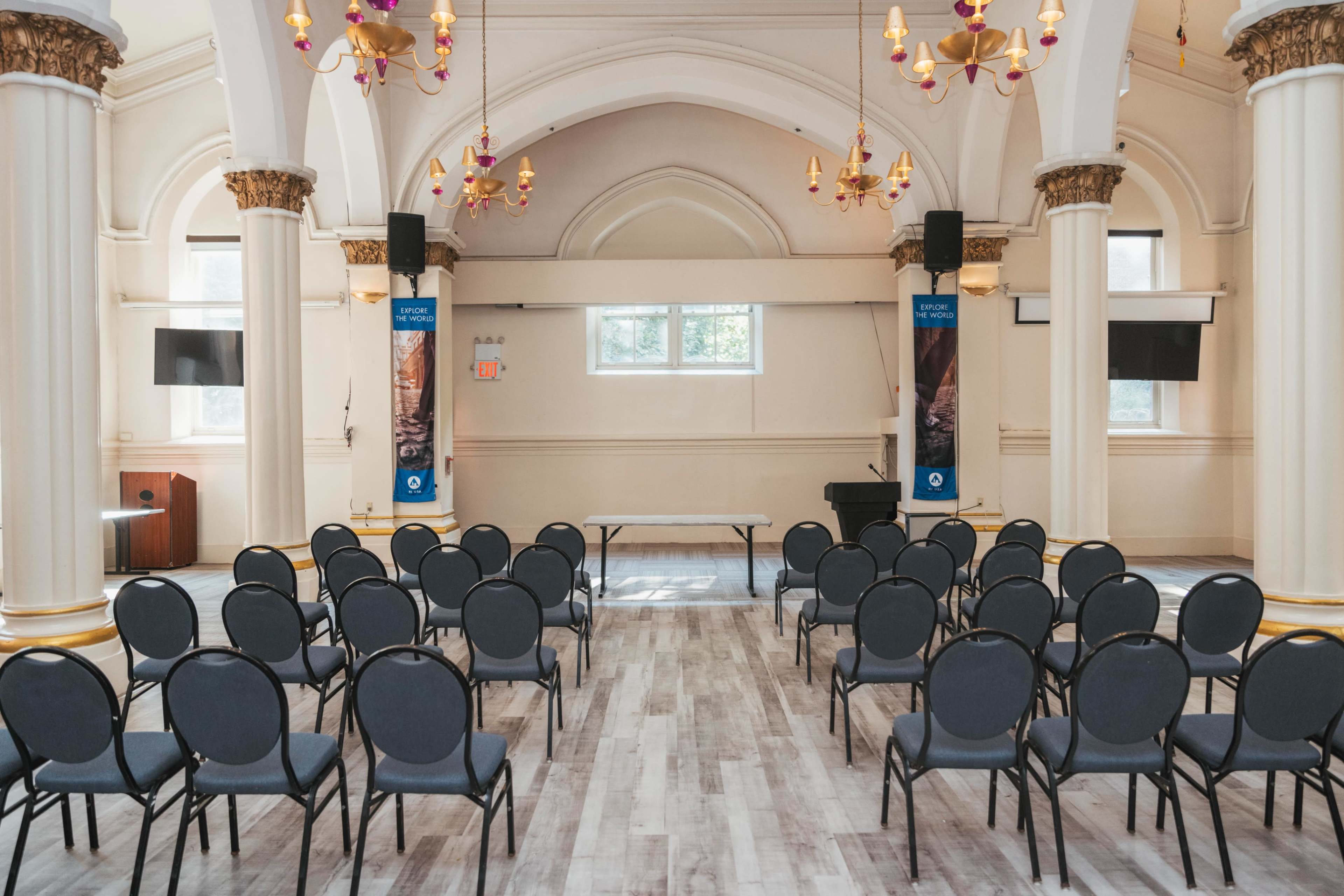 The image shows a spacious event room set up with rows of black chairs facing a small stage, adorned with elegant chandeliers and large pillars.