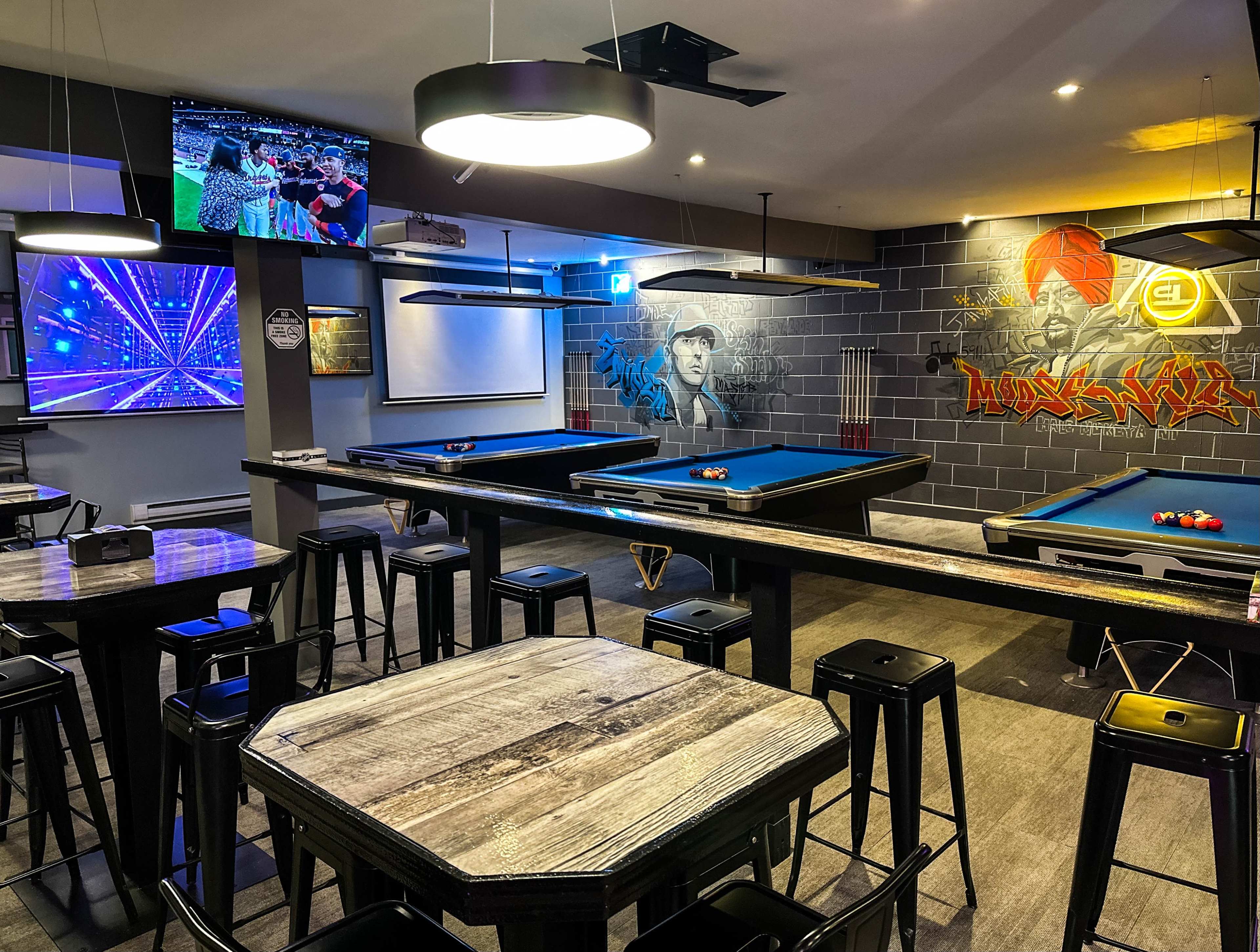 A billiard room featuring several pool tables, bar stools, and a large screen displaying visuals, with wall murals of figures and vibrant artwork.