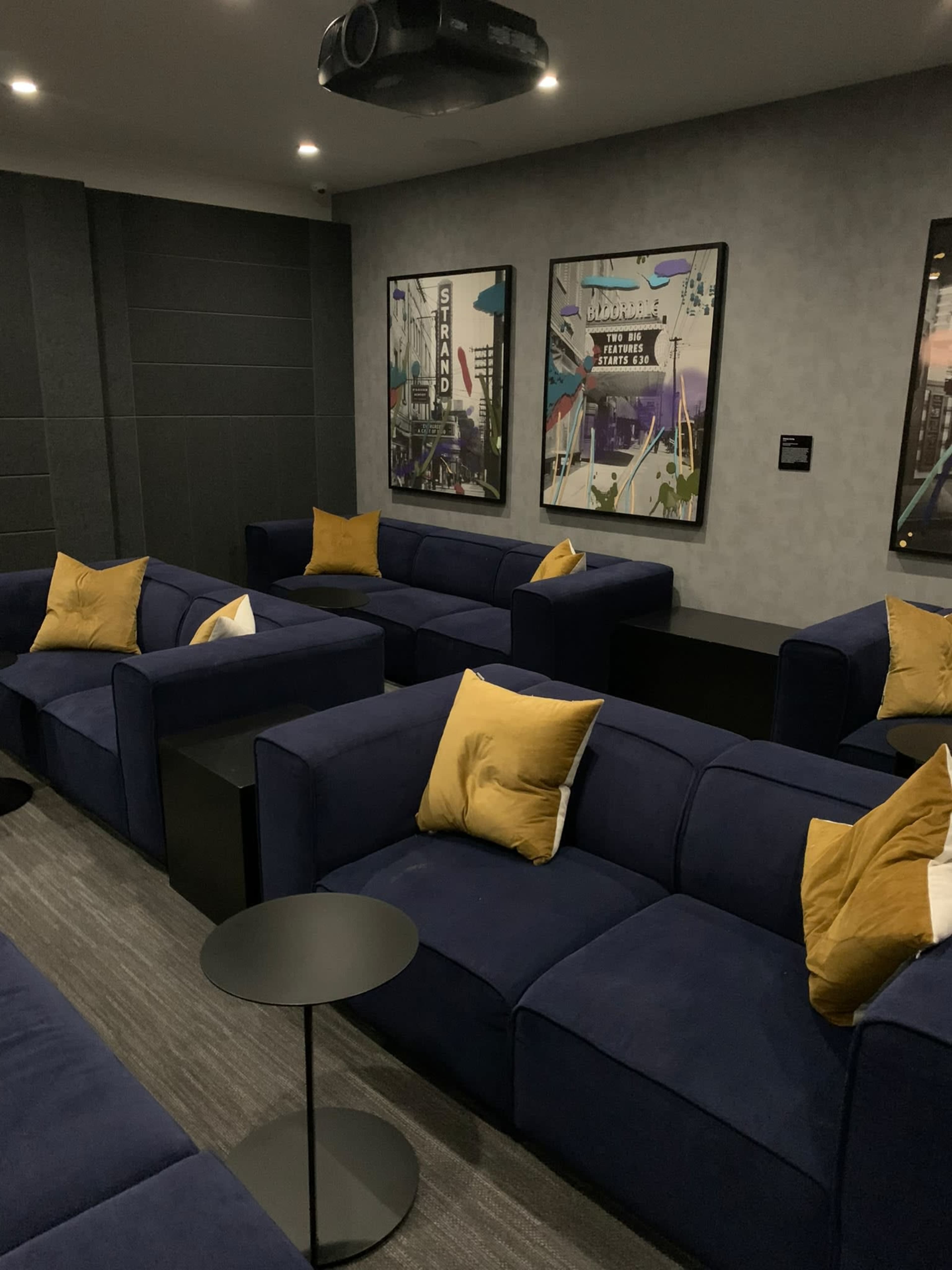 The image shows a modern lounge area with navy blue couches, yellow accent pillows, and two framed artworks on the wall, set against a gray backdrop.