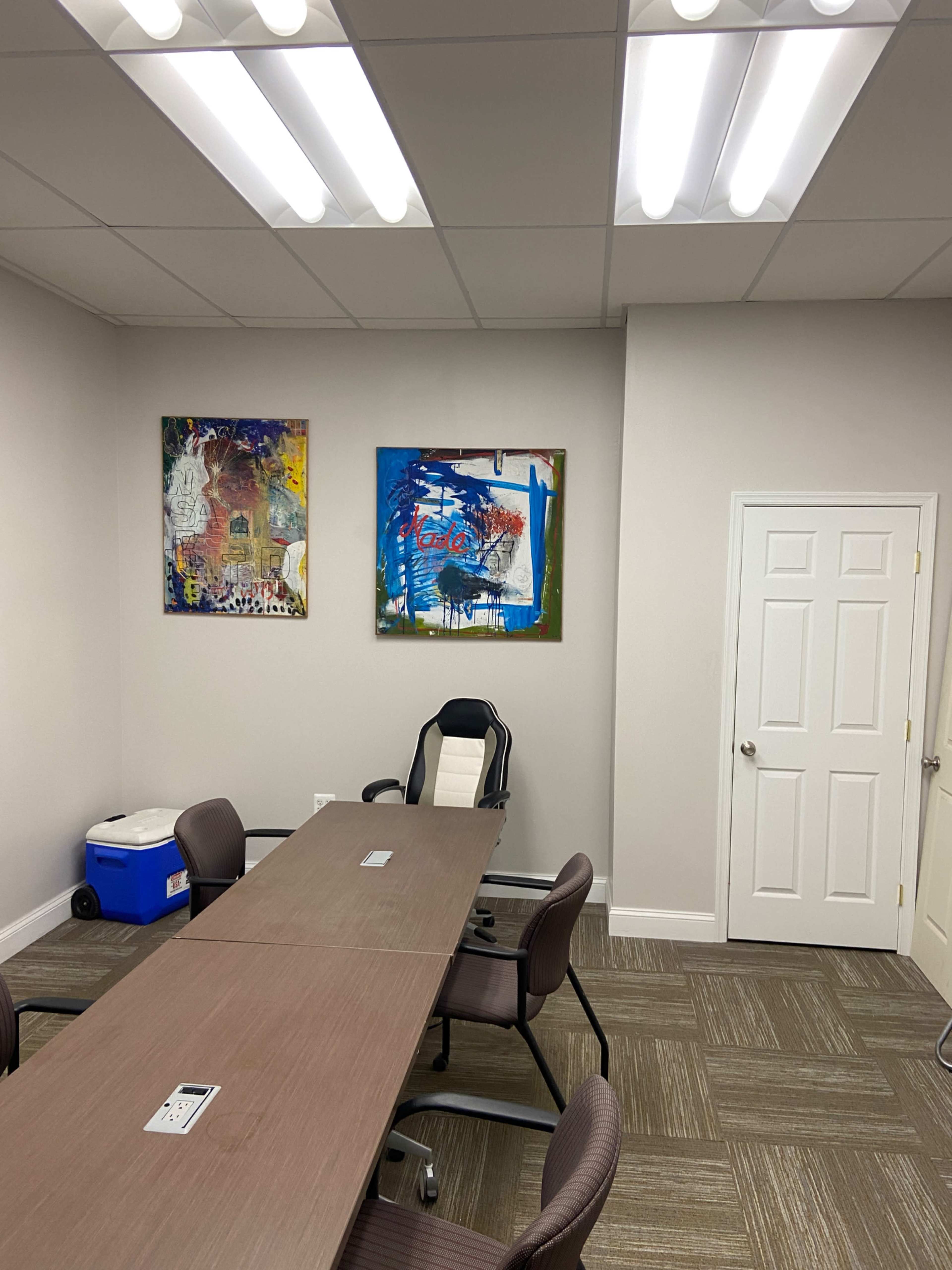 An office conference room features a long table surrounded by chairs, with two colorful abstract paintings on the wall and a door leading to another space.