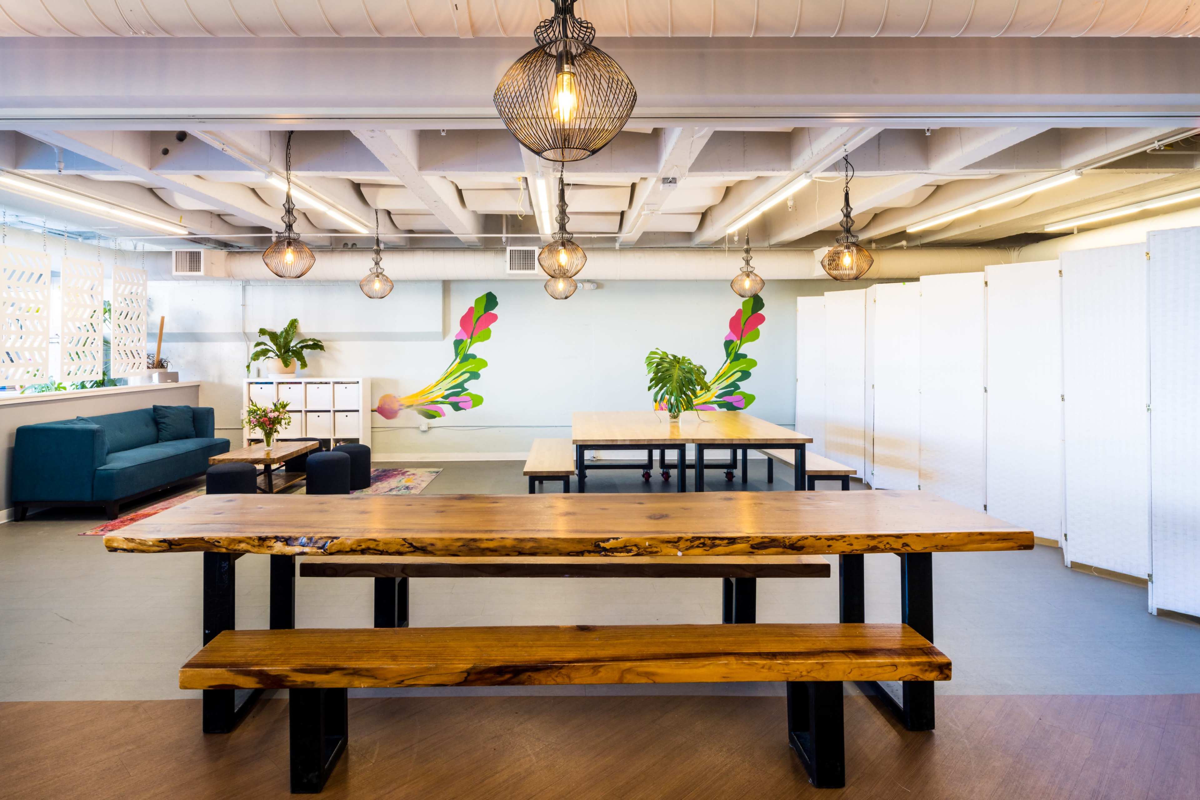 A modern open workspace featuring a long wooden table with benches, a green sofa, and a colorful mural on the wall, illuminated by pendant lights.