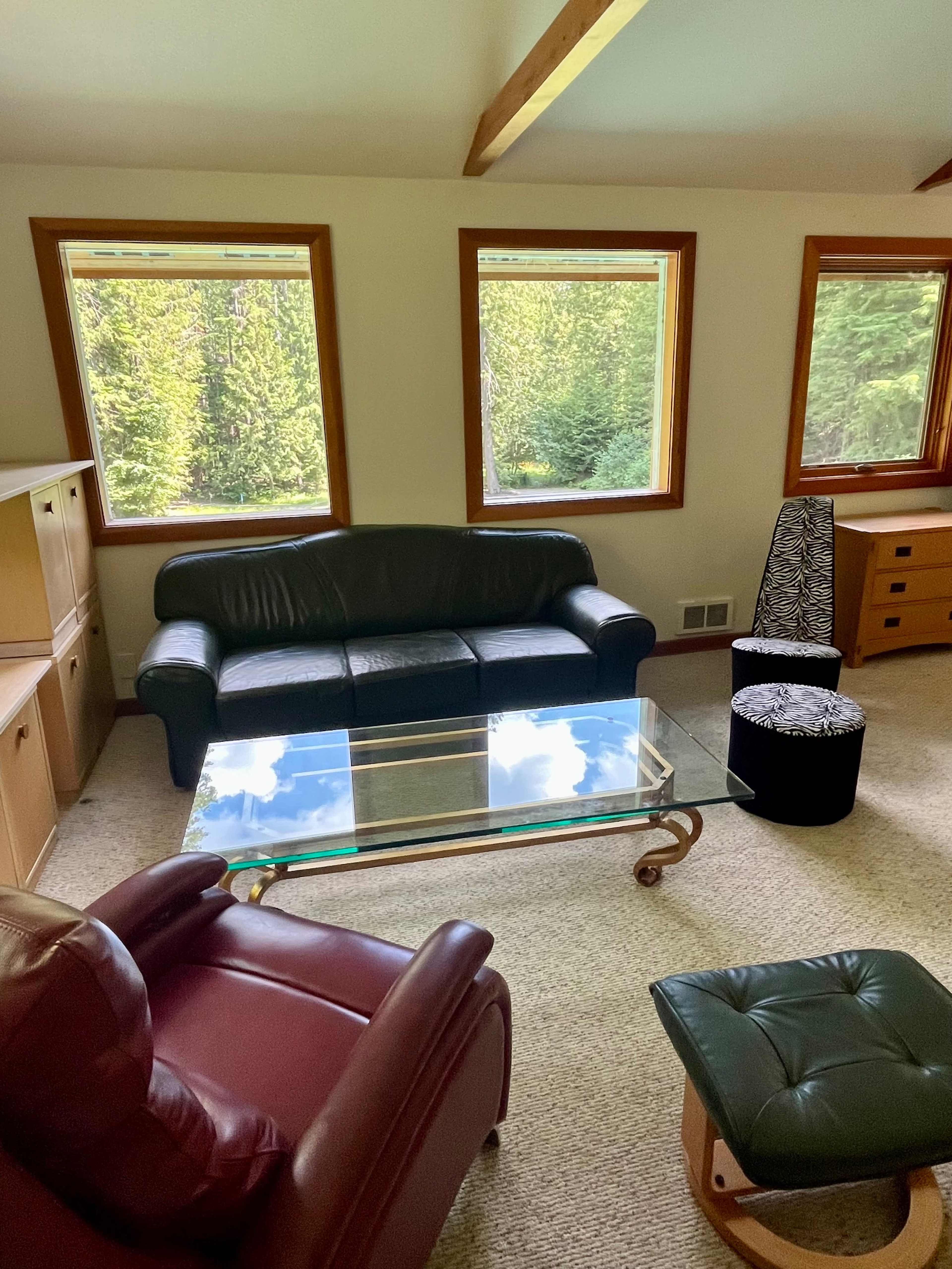 A living room with a black leather sofa, a glass coffee table, a dark ottoman, and wooden furniture arranged near large windows that overlook a forested area.