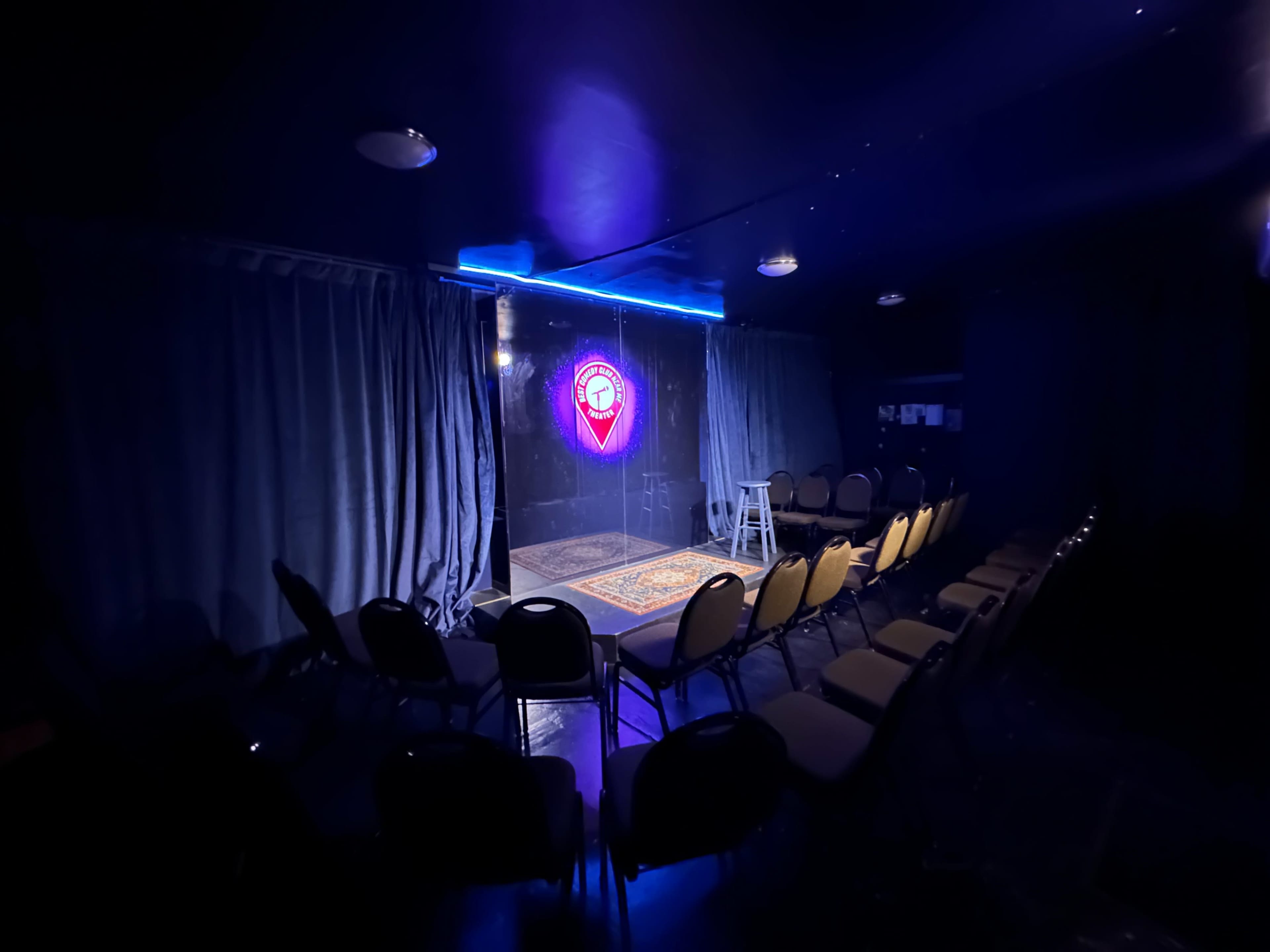The image shows a small performance stage with a neon-lit logo, surrounded by empty chairs arranged in rows.