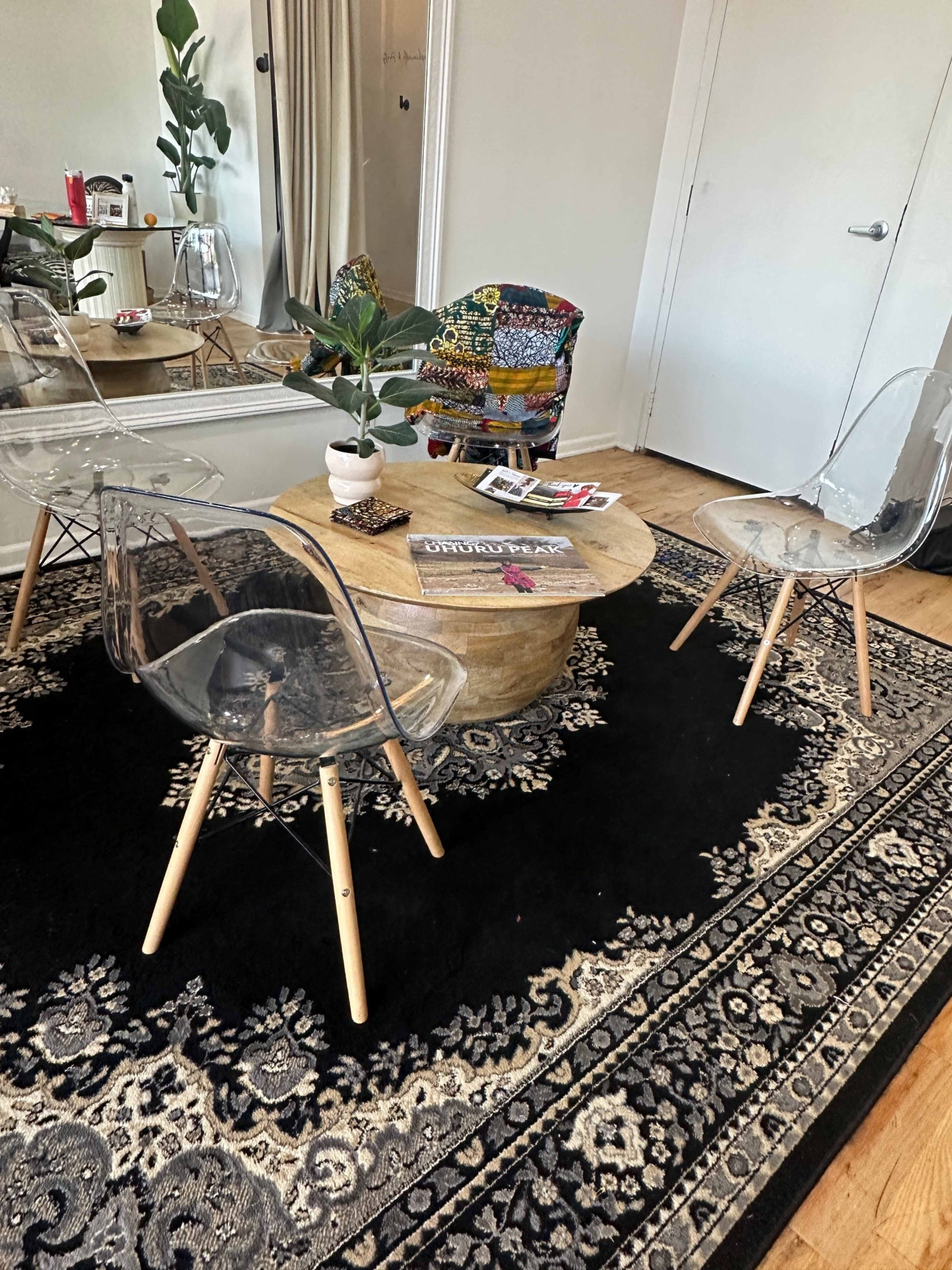 A modern seating area features two transparent chairs and a round wooden coffee table, placed on a patterned black rug.