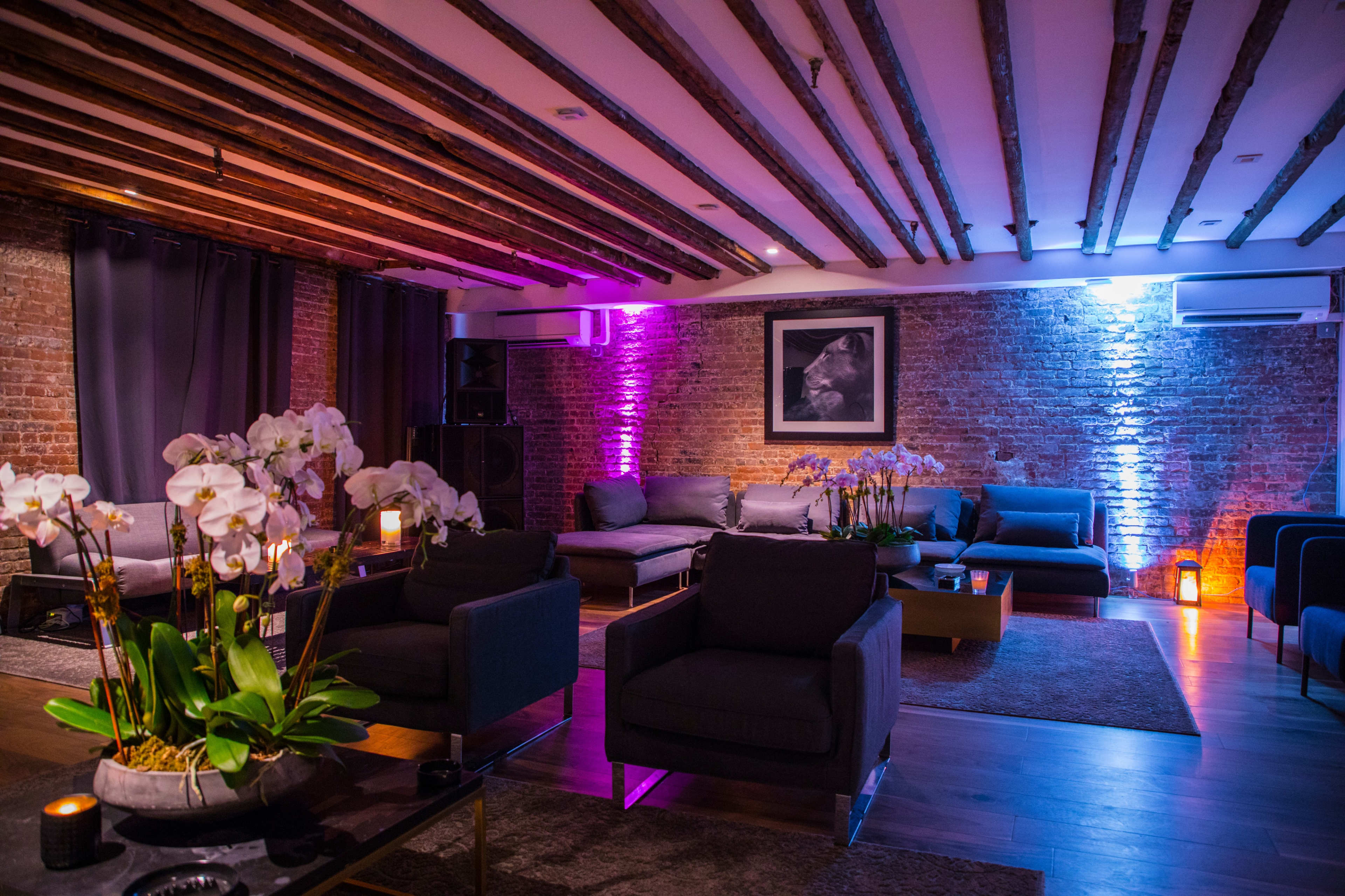 A modern lounge with exposed wooden beams, featuring cozy seating arrangements, decorative orchids, and ambient lighting against a brick wall.