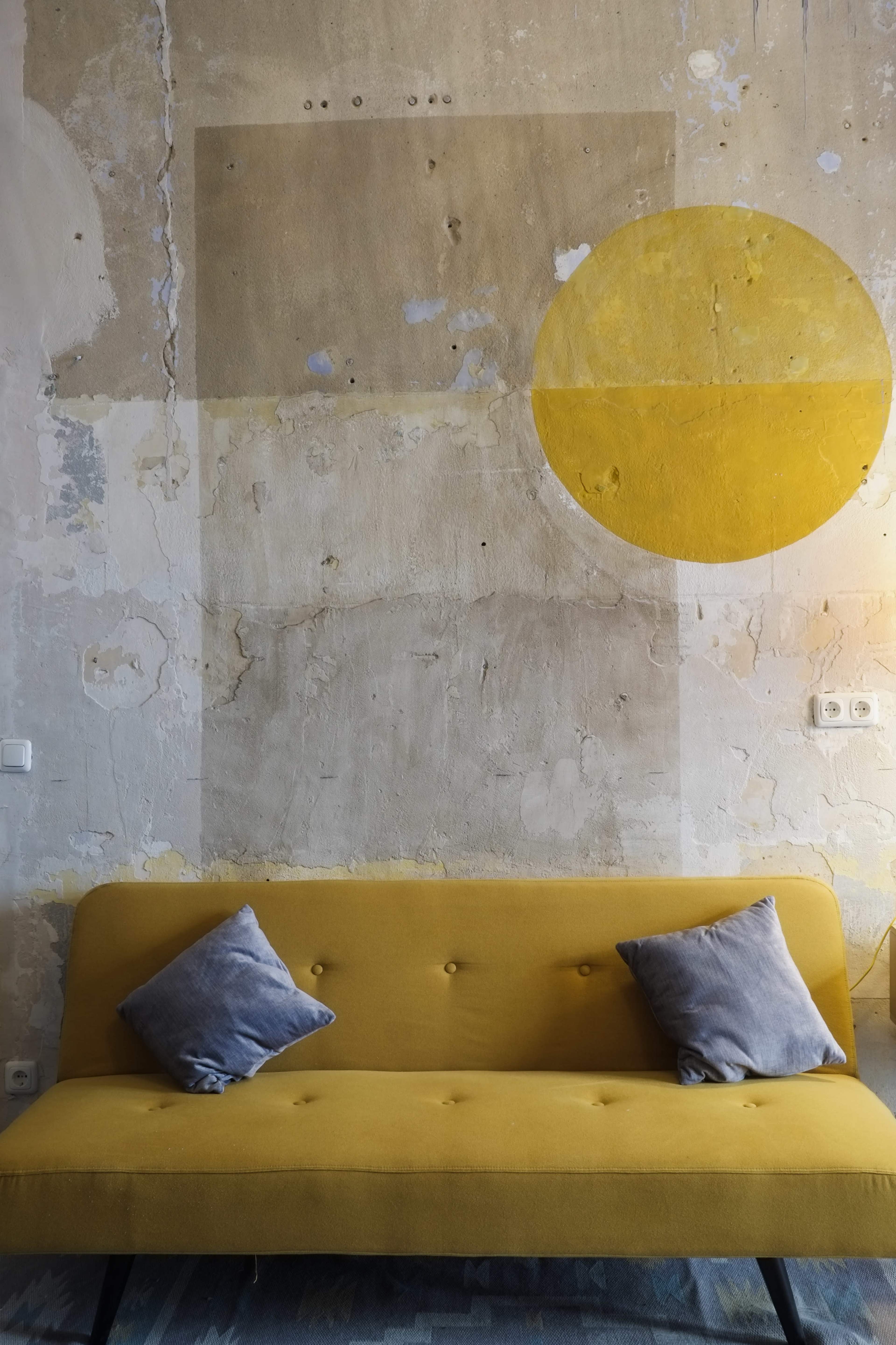 A yellow sofa with two gray pillows is positioned against a weathered wall featuring a large yellow circle and abstract shapes.