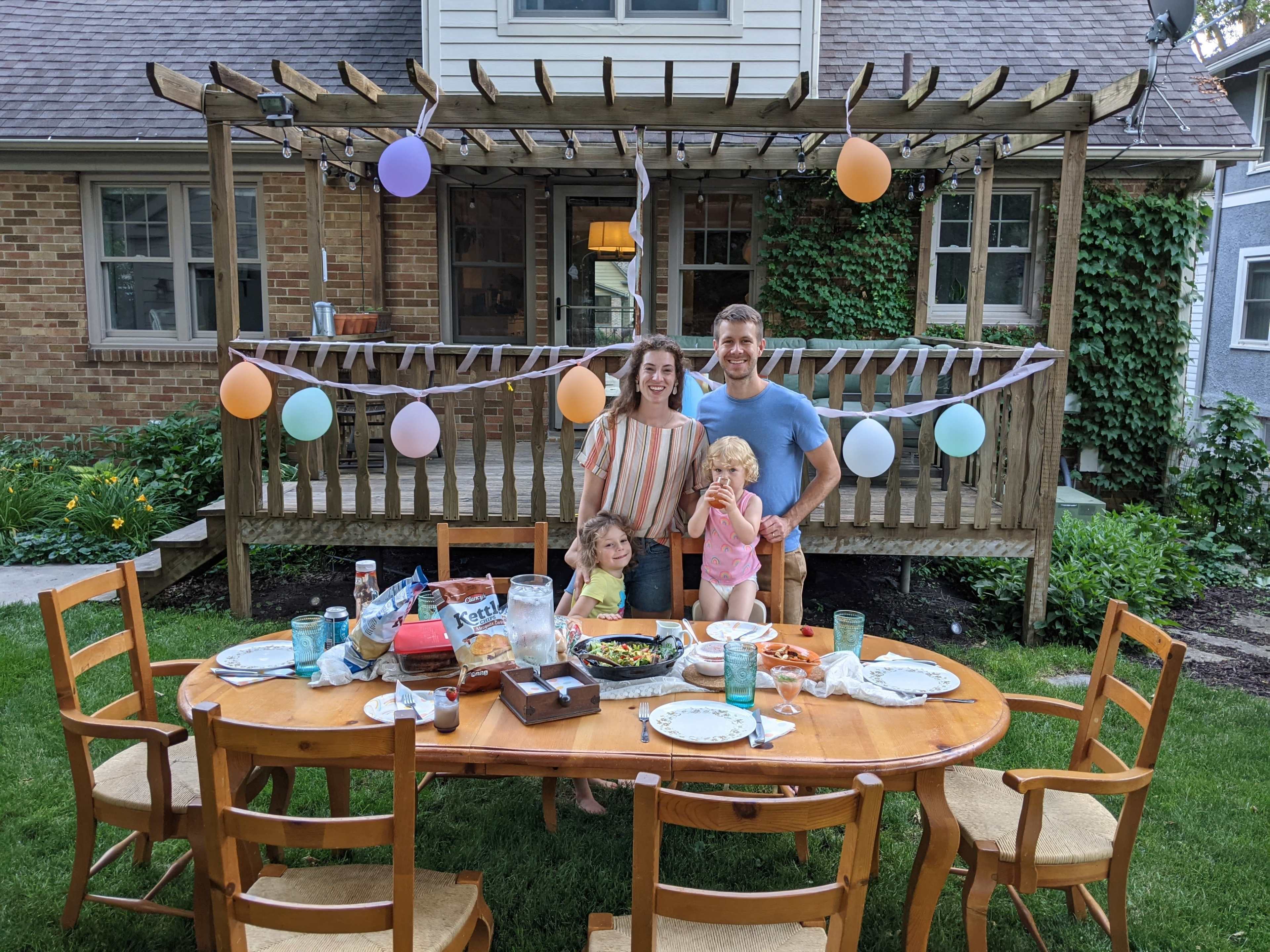 A family poses together in a backyard setting with a decorated table set for a meal, surrounded by greenery and balloons.