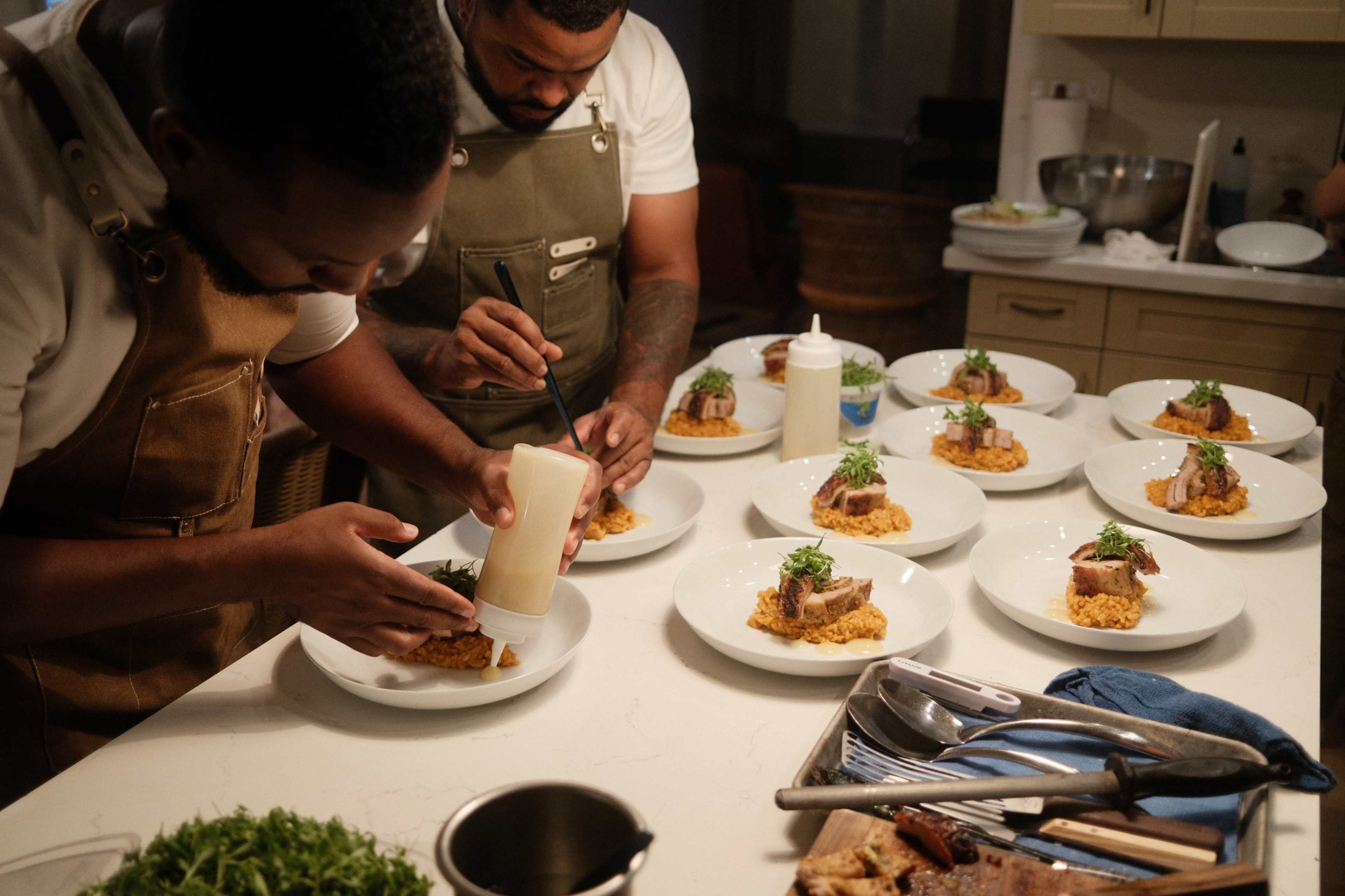 Two chefs in aprons plate food on white dishes in a kitchen.