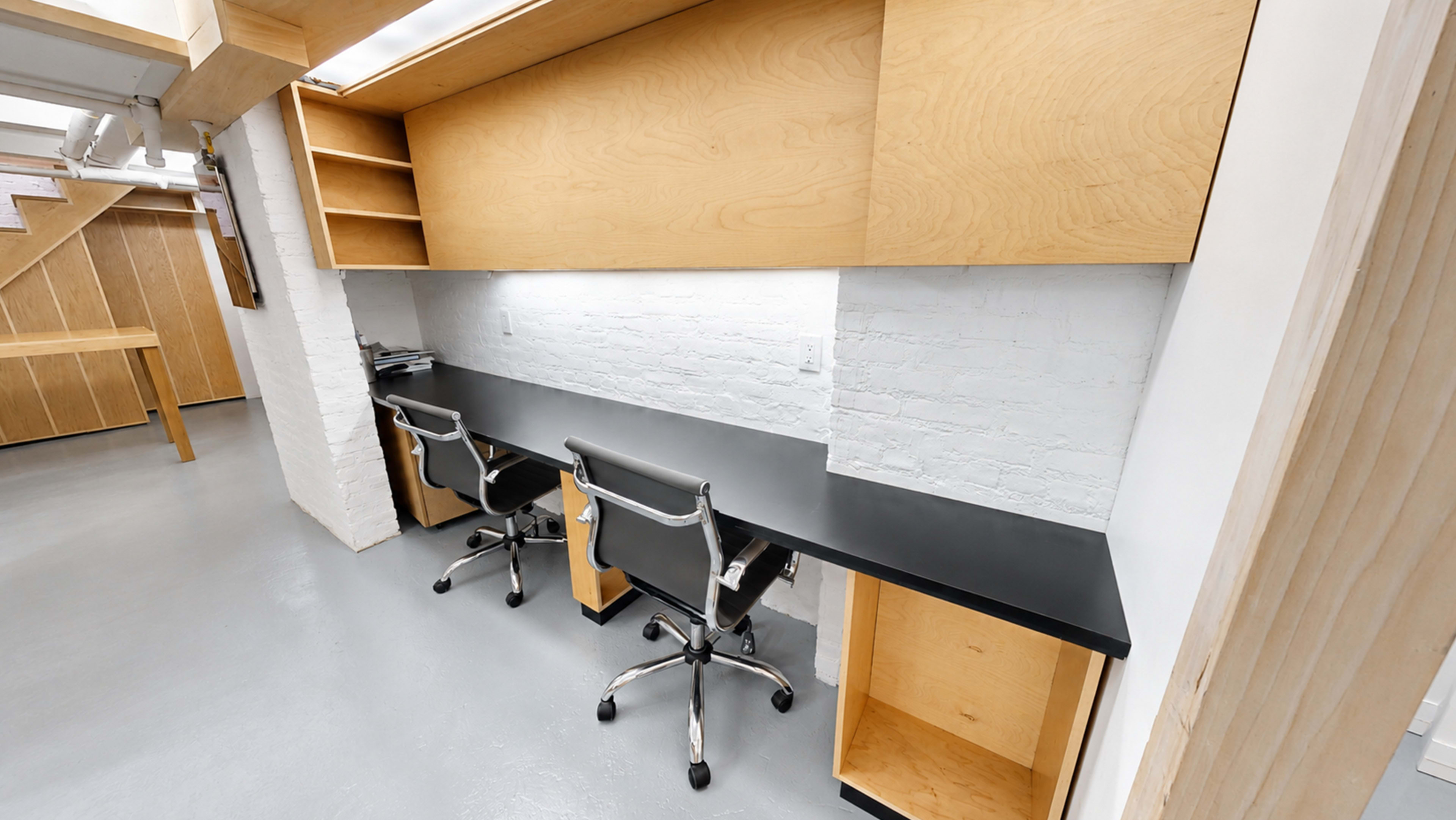 The image shows a minimalistic workspace featuring a long black desk with two ergonomic chairs and wooden shelving above.